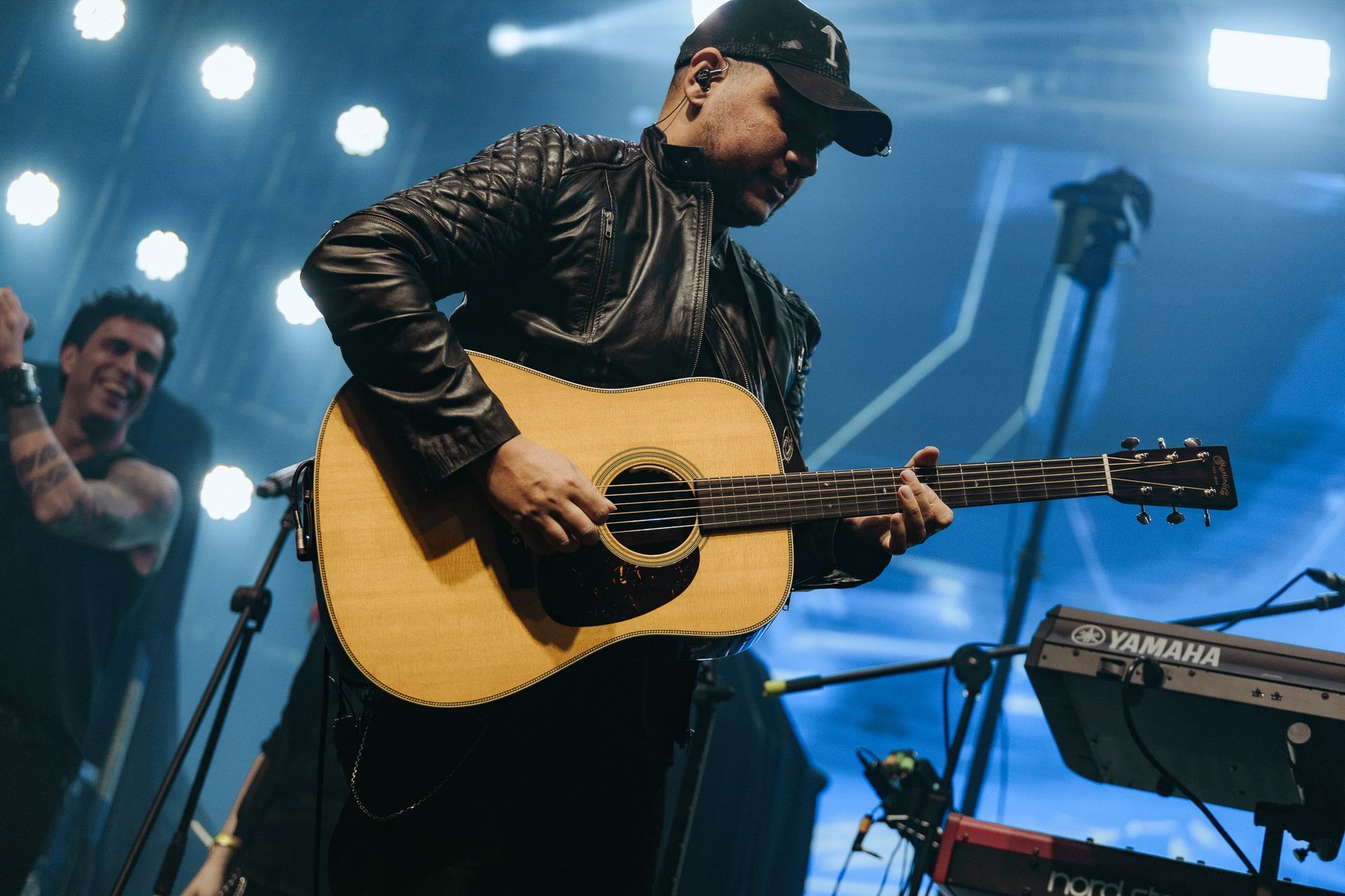 Um artista tocando violão no palco, vestindo jaqueta preta e boné de beisebol, com outro homem ao fundo.