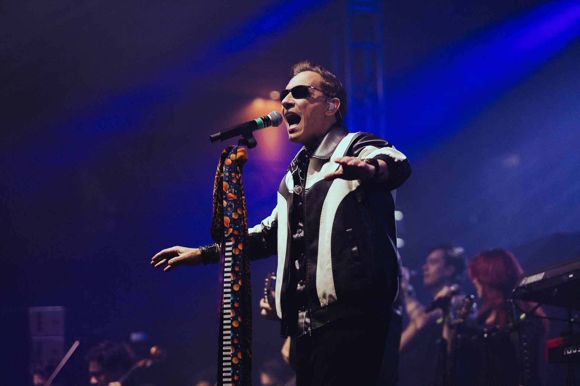Um artista cantando ao microfone no palco, vestindo uma jaqueta preta e branca. Luzes azuis no palco.