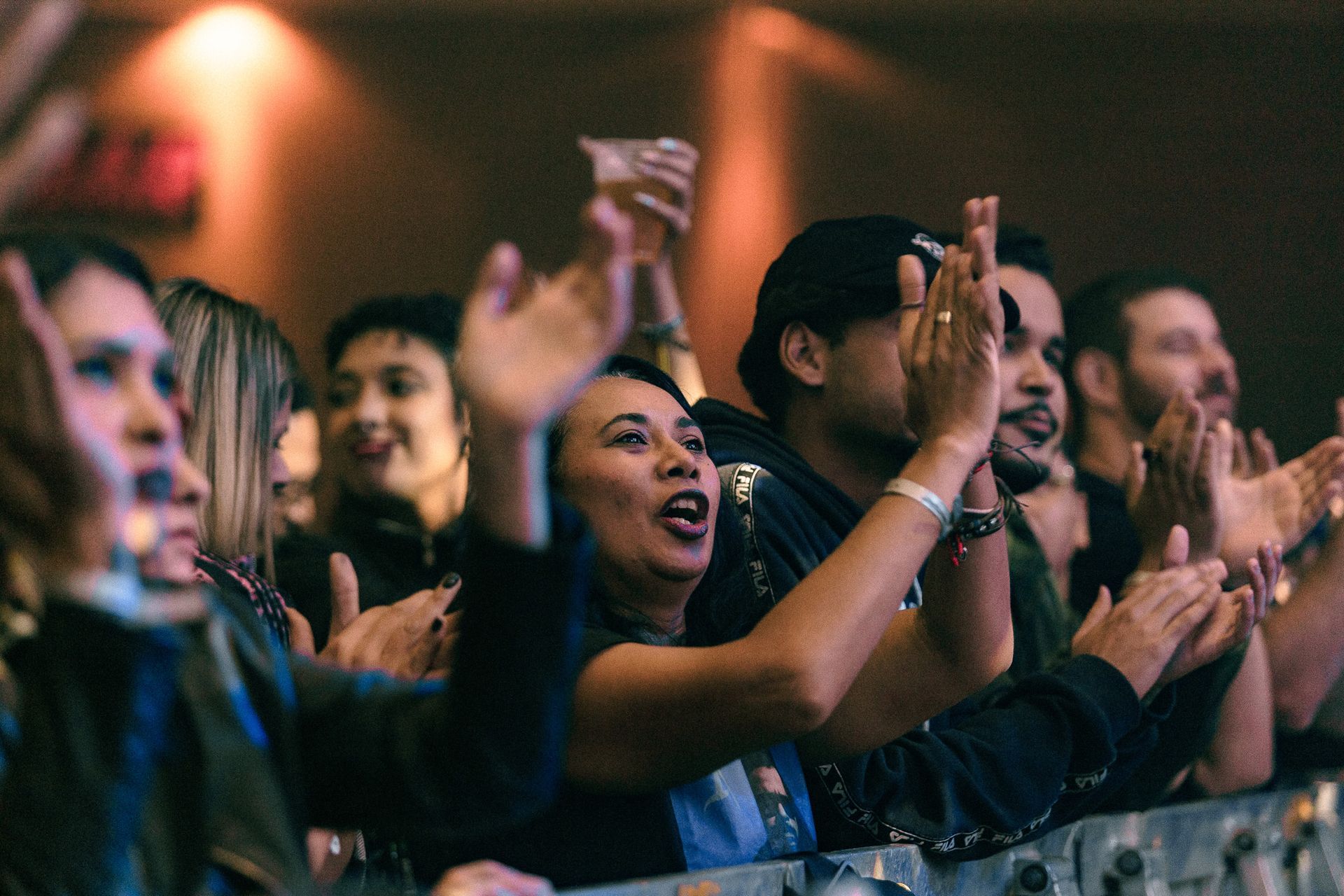 Público aplaudindo em um show, alguns com a boca aberta. Iluminação de palco aconchegante.