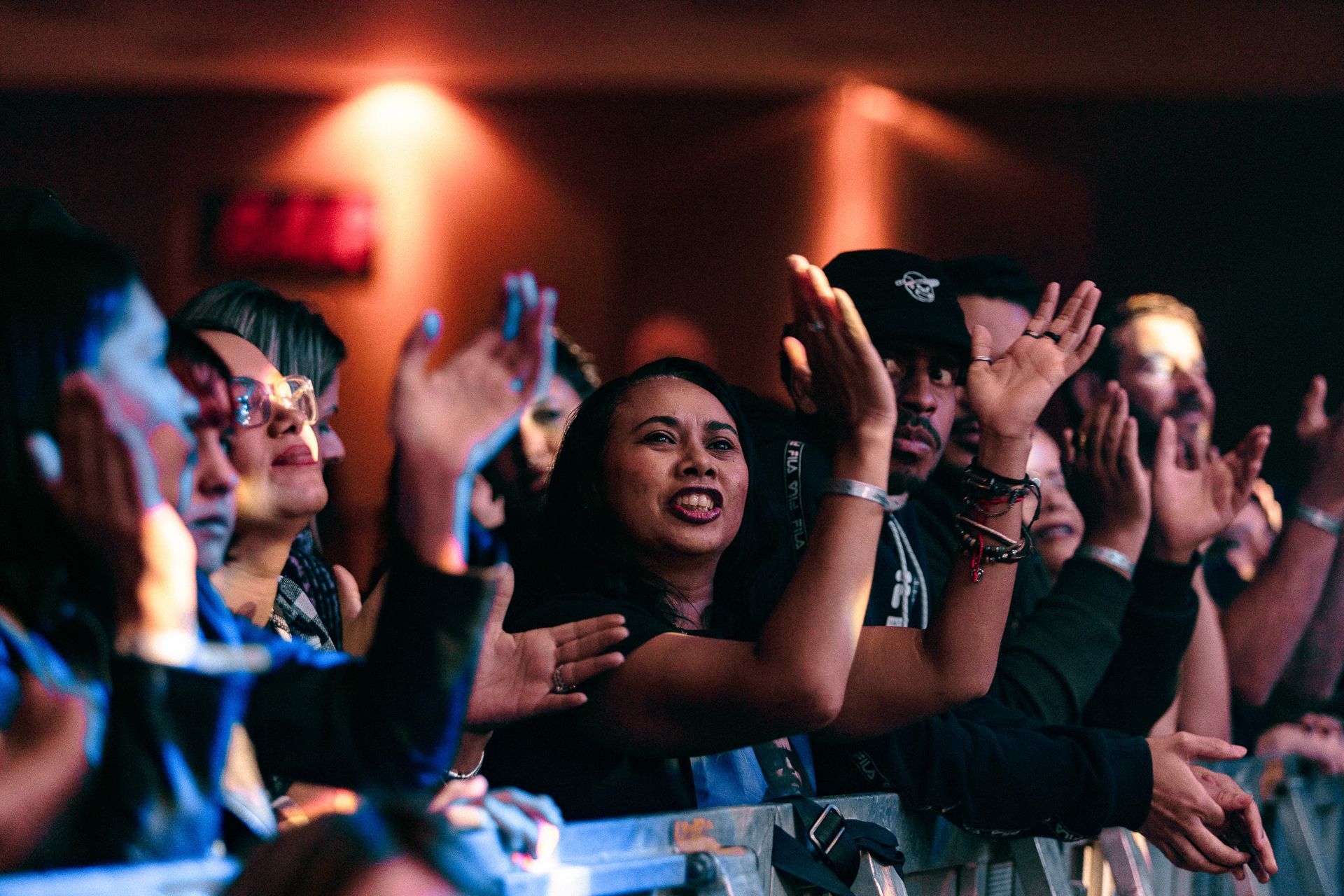 Pessoas com os braços erguidos, algumas aplaudindo, em um show. Luzes do palco iluminam a multidão.