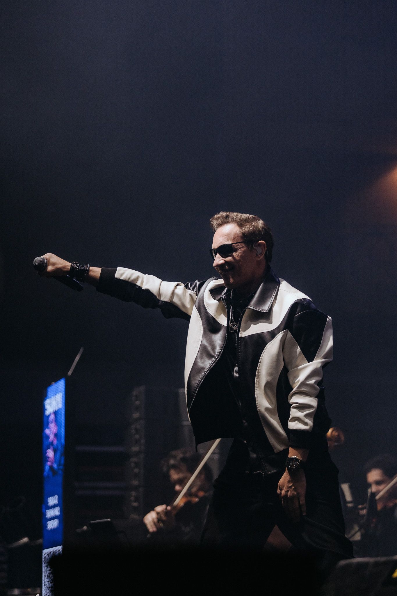 Um artista no palco, vestindo jaqueta branca e preta, segurando um microfone e estendendo a mão para a plateia.