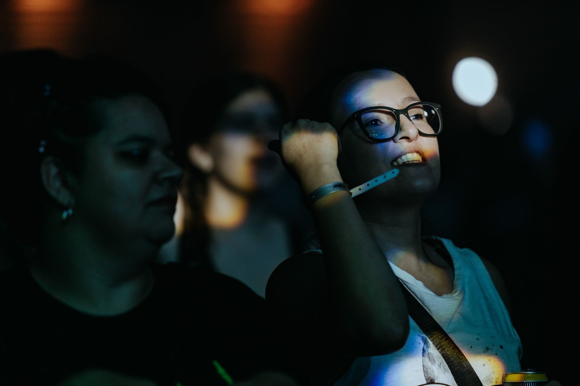 Mulher de óculos em um show, com a mão levantada. Ambiente escuro com luzes de palco. Outras pessoas na plateia observam.