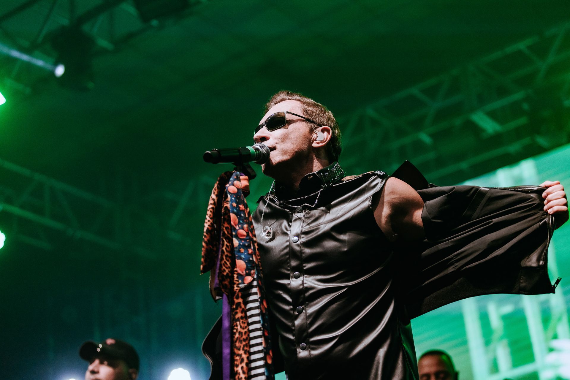 Um artista de óculos escuros no palco, segurando um microfone, vestindo camisa preta e capa, iluminação verde.