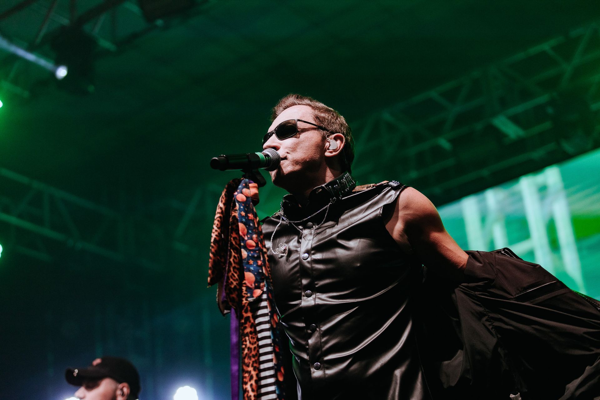 Um artista de óculos escuros cantando em um microfone no palco, vestindo um colete de couro preto e segurando um lenço estampado.