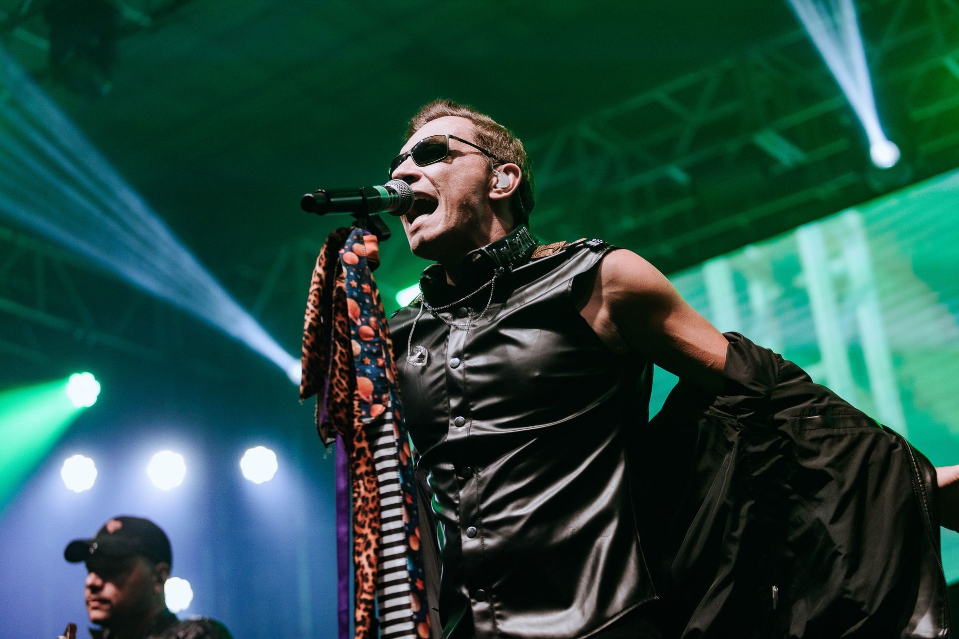 Cantor no palco, jaqueta de couro, óculos escuros, cantando ao microfone. Iluminação verde no palco.