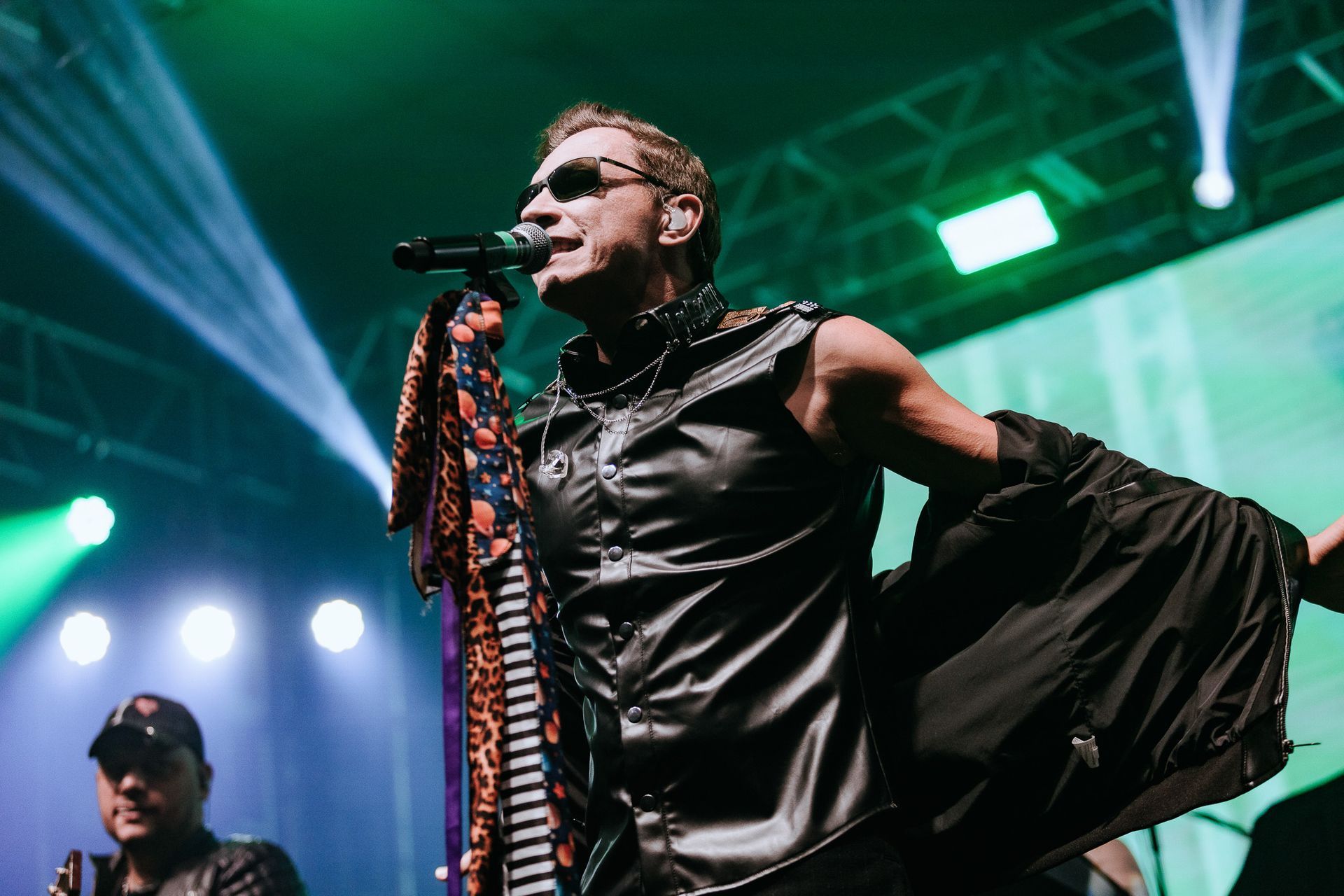 Um artista de óculos escuros cantando ao microfone no palco, colete de couro preto, luzes ao fundo.