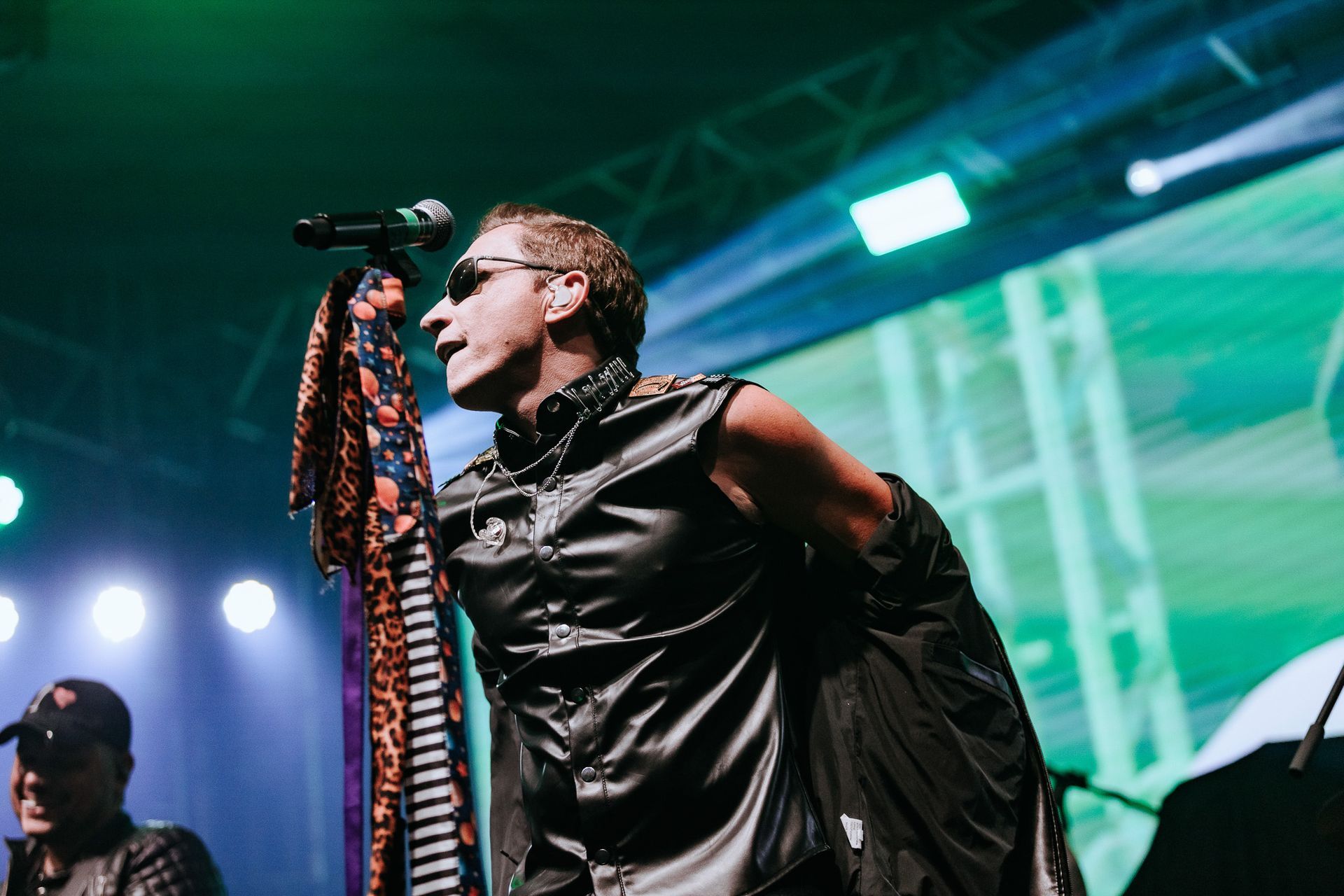 Um cantor de colete preto e óculos escuros se apresenta no palco, com o microfone na mão.