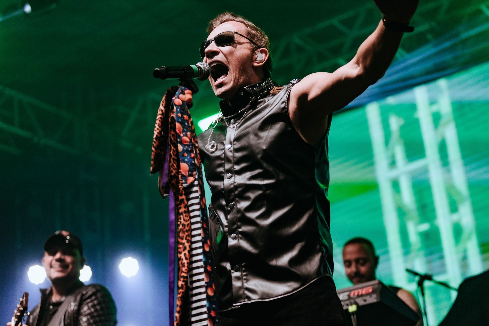 Um artista cantando com paixão no palco, vestindo um colete preto e óculos escuros, com os membros da banda ao fundo.