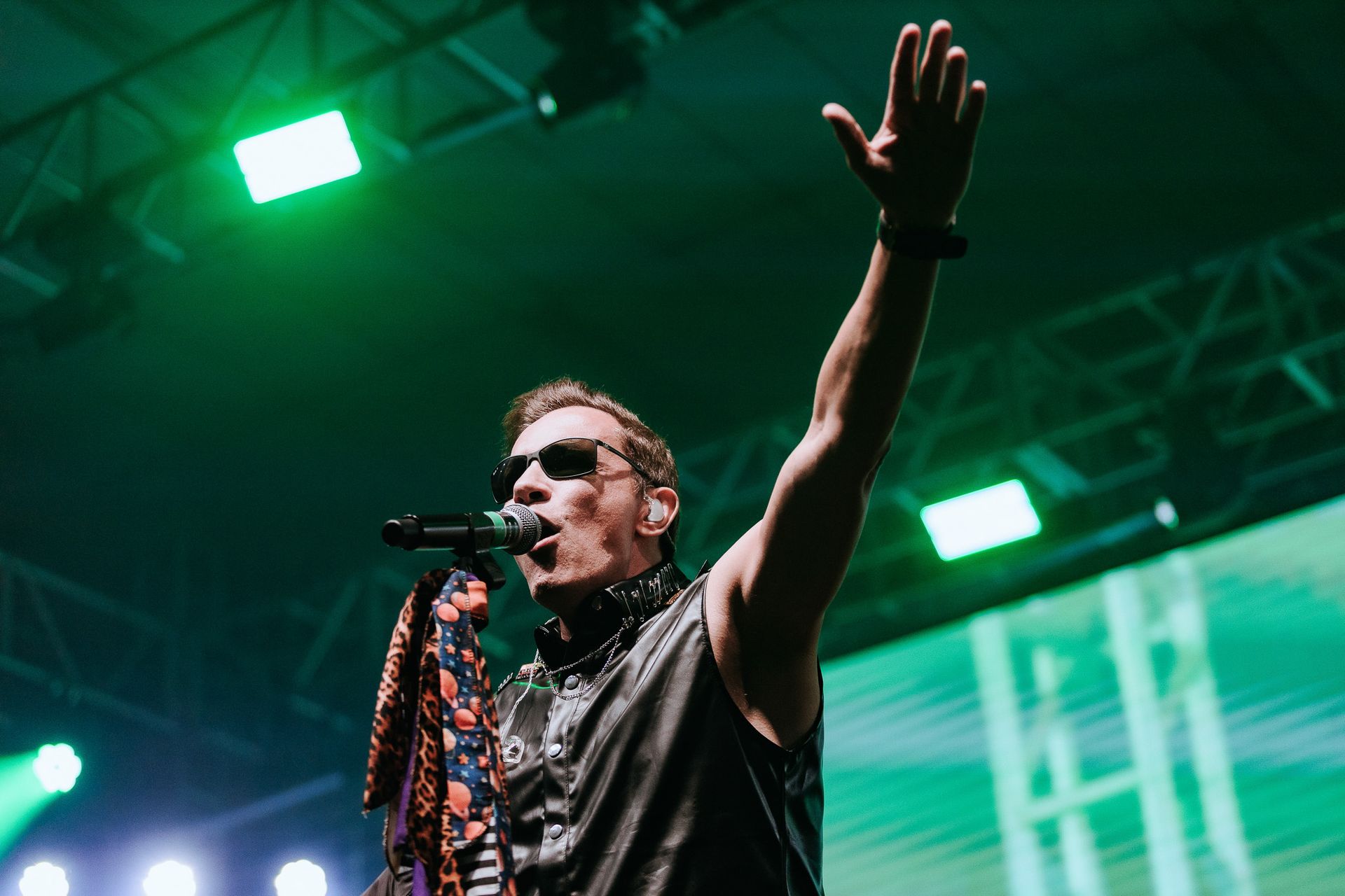 Um homem de óculos escuros canta em um microfone, levantando o braço em uma apresentação sob luzes verdes de palco.
