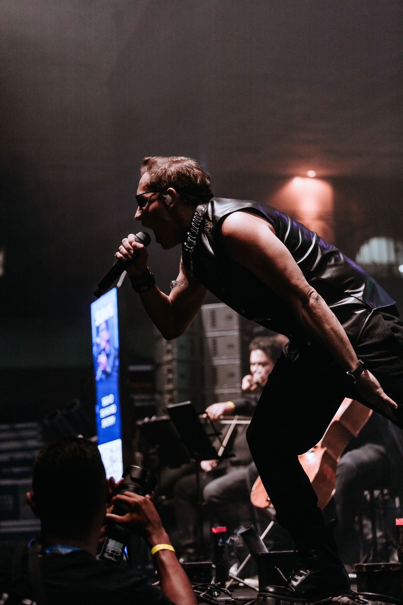 Cantor no palco, cantando ao microfone. Roupas escuras, luzes de palco, banda ao fundo.