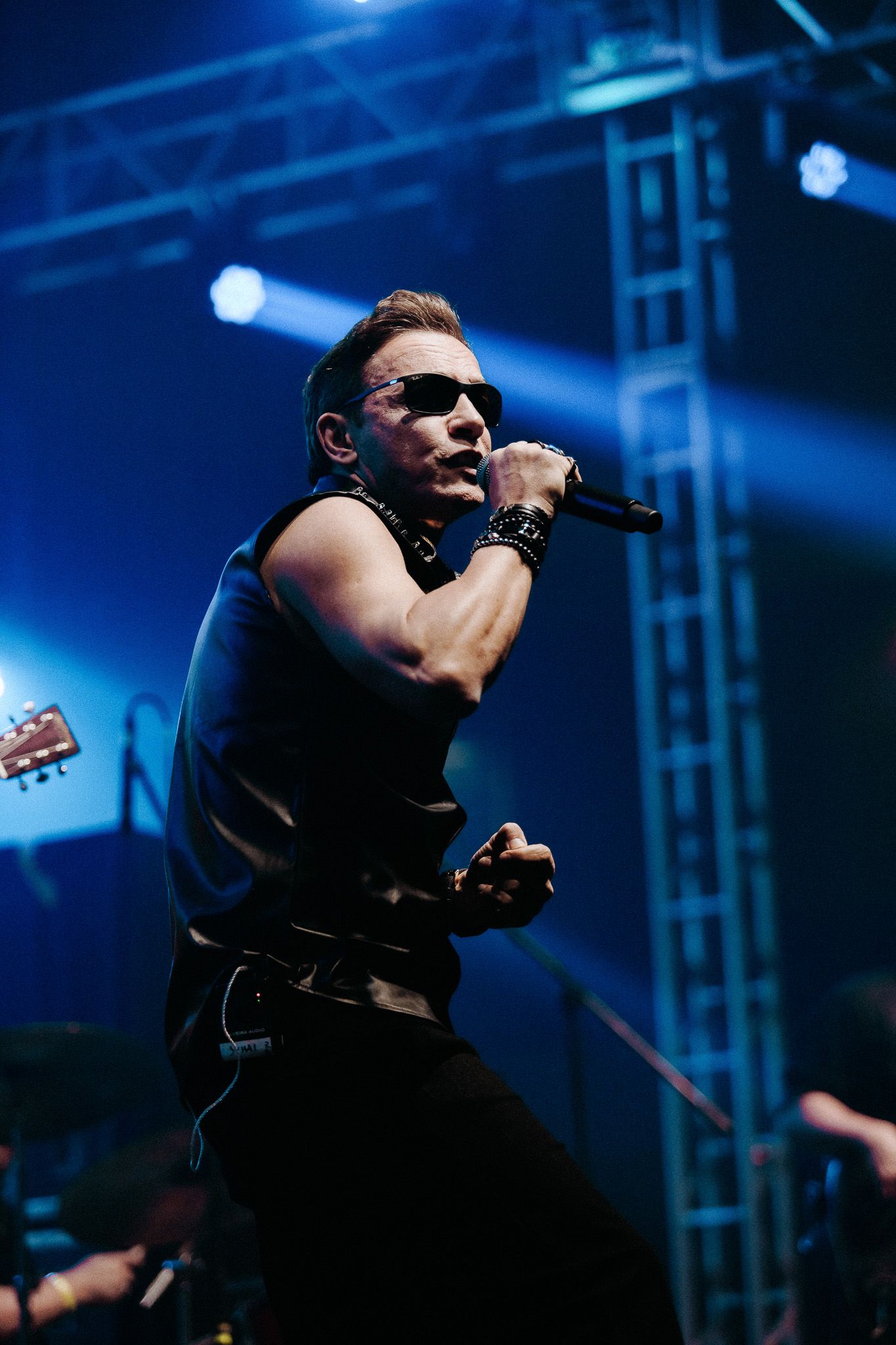 Cantor no palco, usando óculos escuros, colete e calça pretos, segurando um microfone, se apresentando
