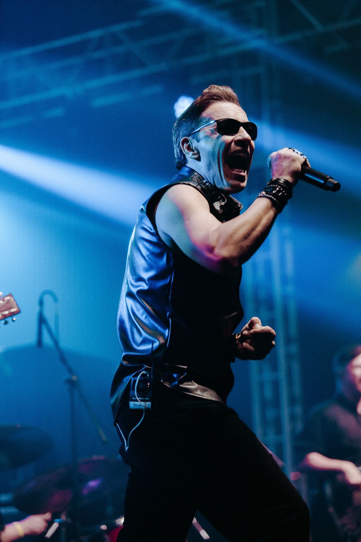 Um cantor de óculos escuros se apresenta no palco, gesticulando com um microfone. O palco está iluminado 