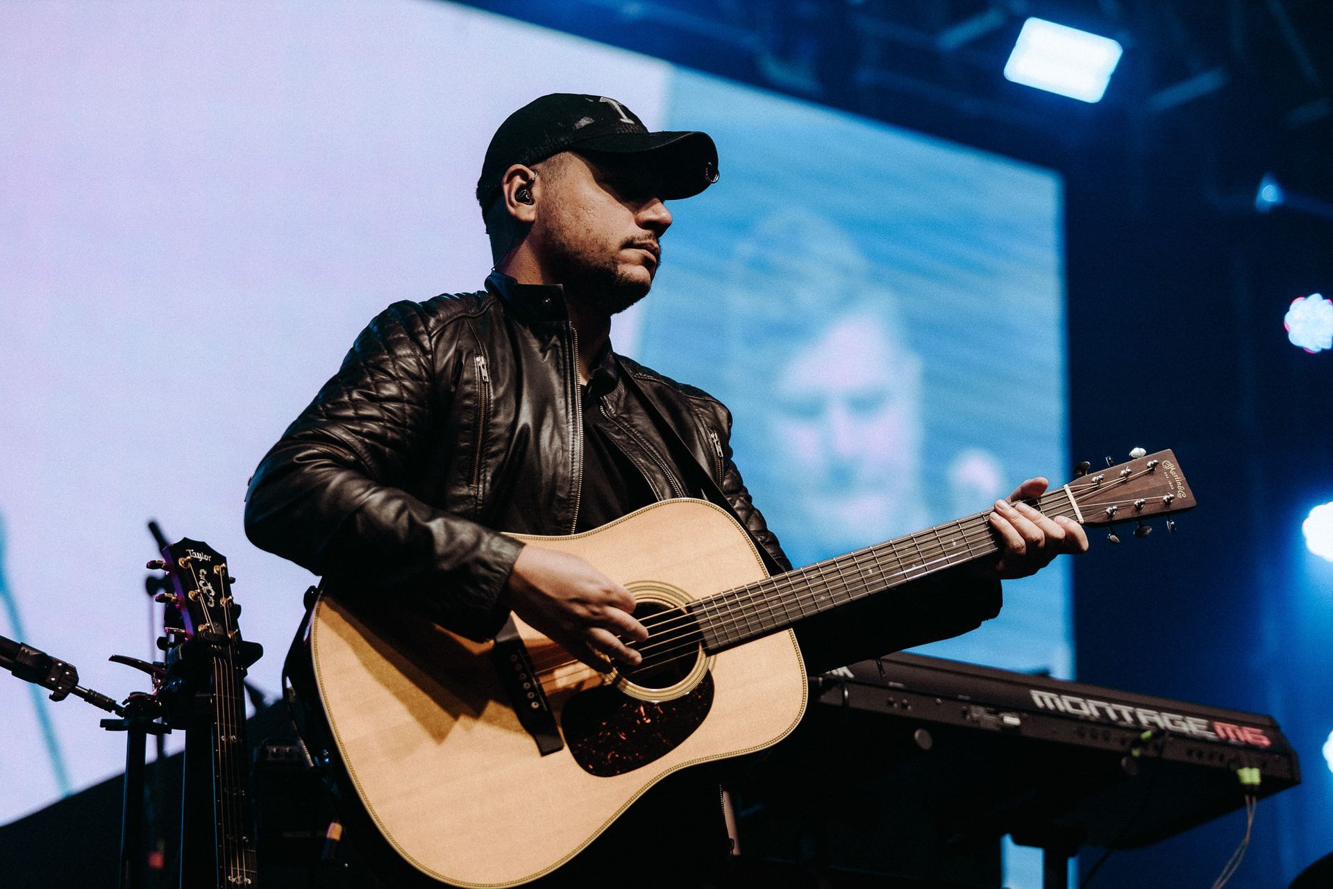 Homem tocando violão no palco, vestindo jaqueta preta e boné de beisebol, com fundo desfocado.