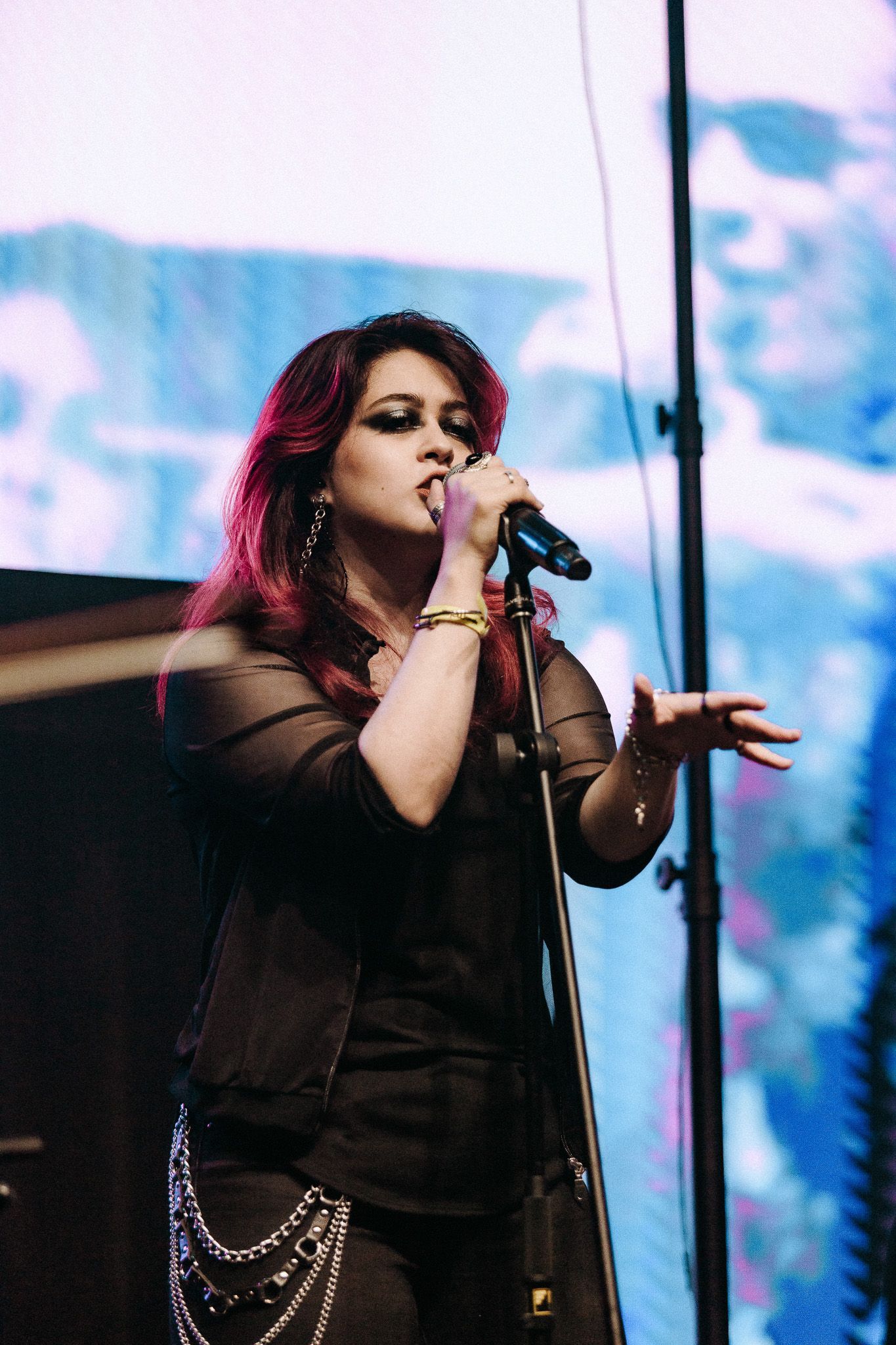Mulher com mechas vermelhas no cabelo canta ao microfone no palco, vestindo uma roupa preta.