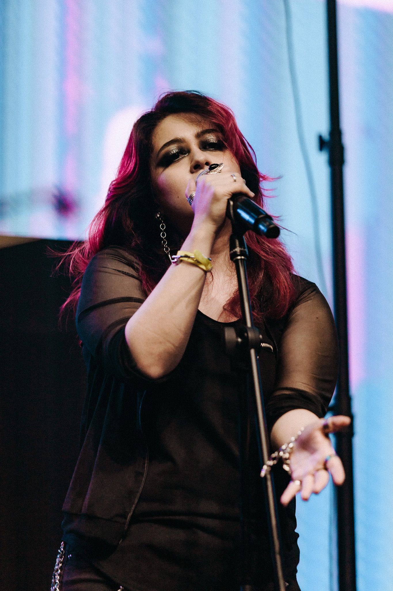 Mulher cantando ao microfone no palco; vestindo roupa preta, cabelo com reflexos vermelhos, mãos estendidas.