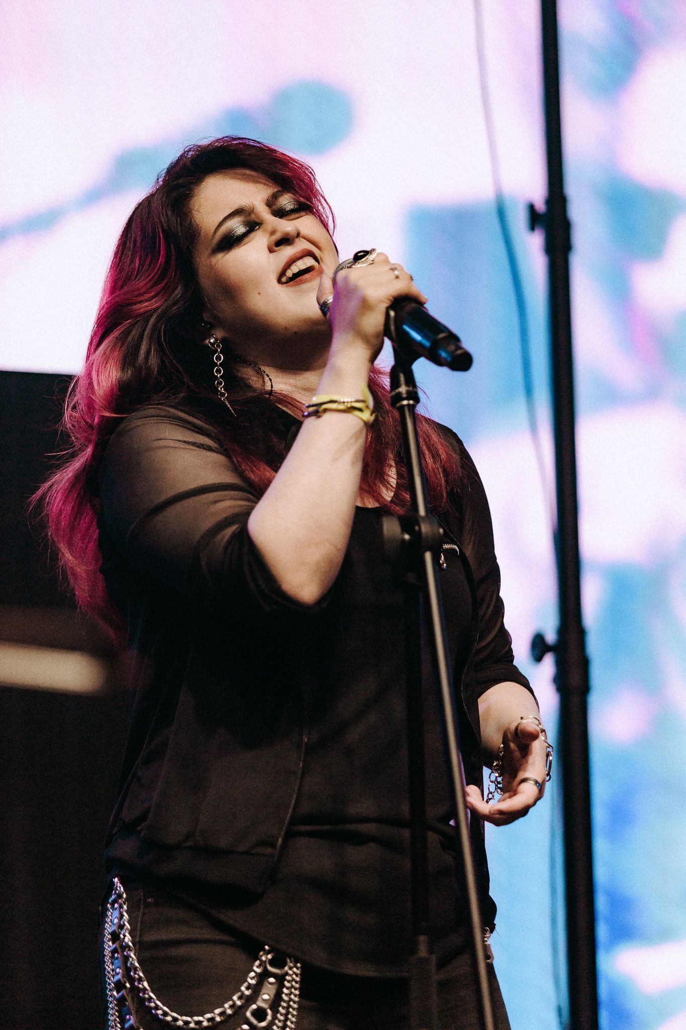 Mulher cantando ao microfone no palco; roupa preta, cabelo ruivo, iluminação de palco.