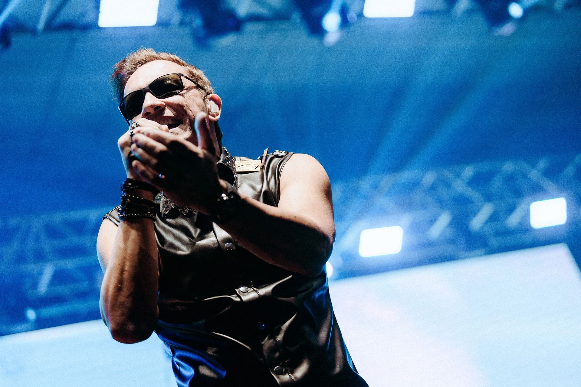 Um homem de óculos escuros e colete de couro cantando no palco, com as mãos perto da boca. Iluminação azul.