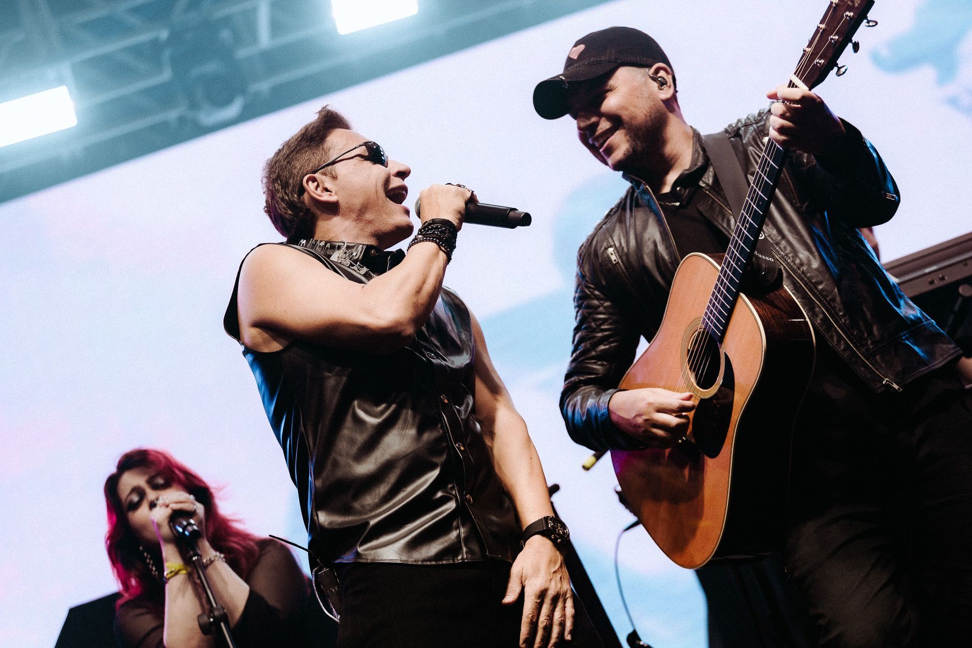 Uma banda se apresentando no palco: um vocalista com um microfone, um guitarrista e uma backing vocal.