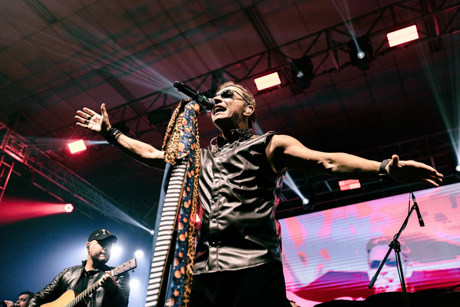 Cantor no palco com os braços estendidos e microfone na mão. Banda ao fundo, luzes de palco.