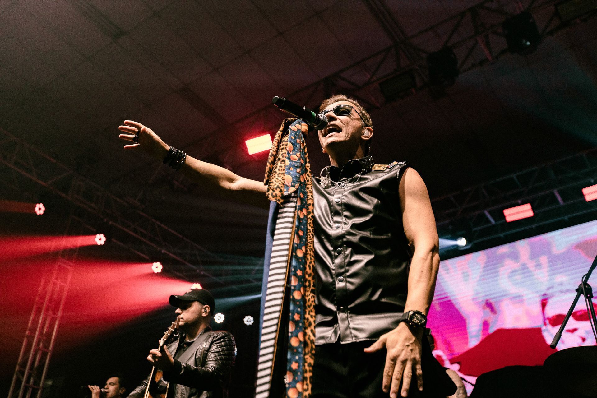 Um artista de óculos escuros e jaqueta aberta no palco, com o braço levantado, cantando em um microfone.