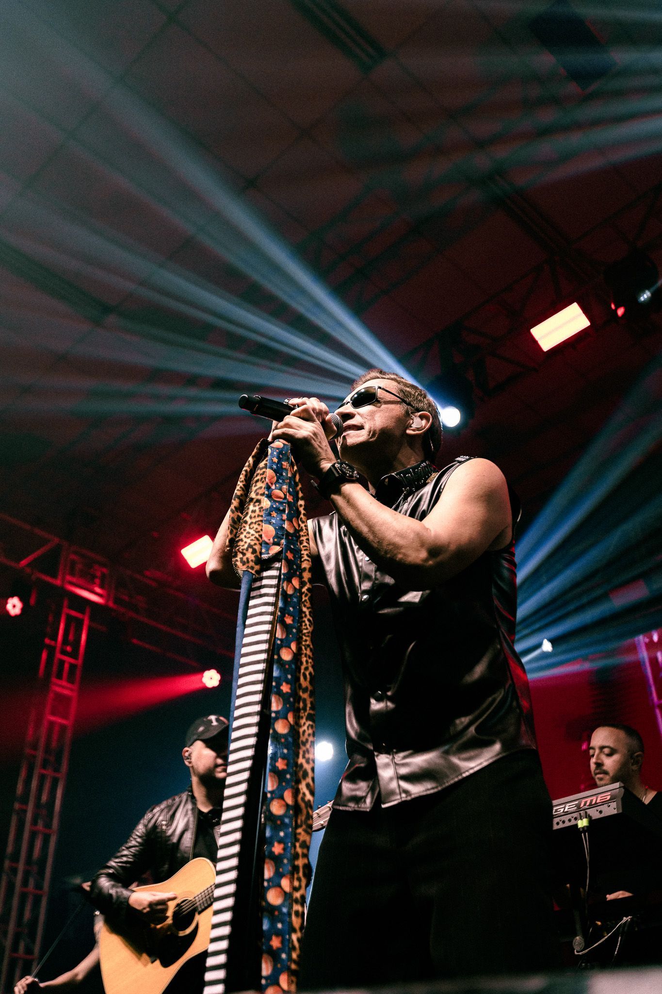 Cantor no palco, segurando o microfone, com seus companheiros de banda. Holofotes e luzes vermelhas iluminam a apresentação.