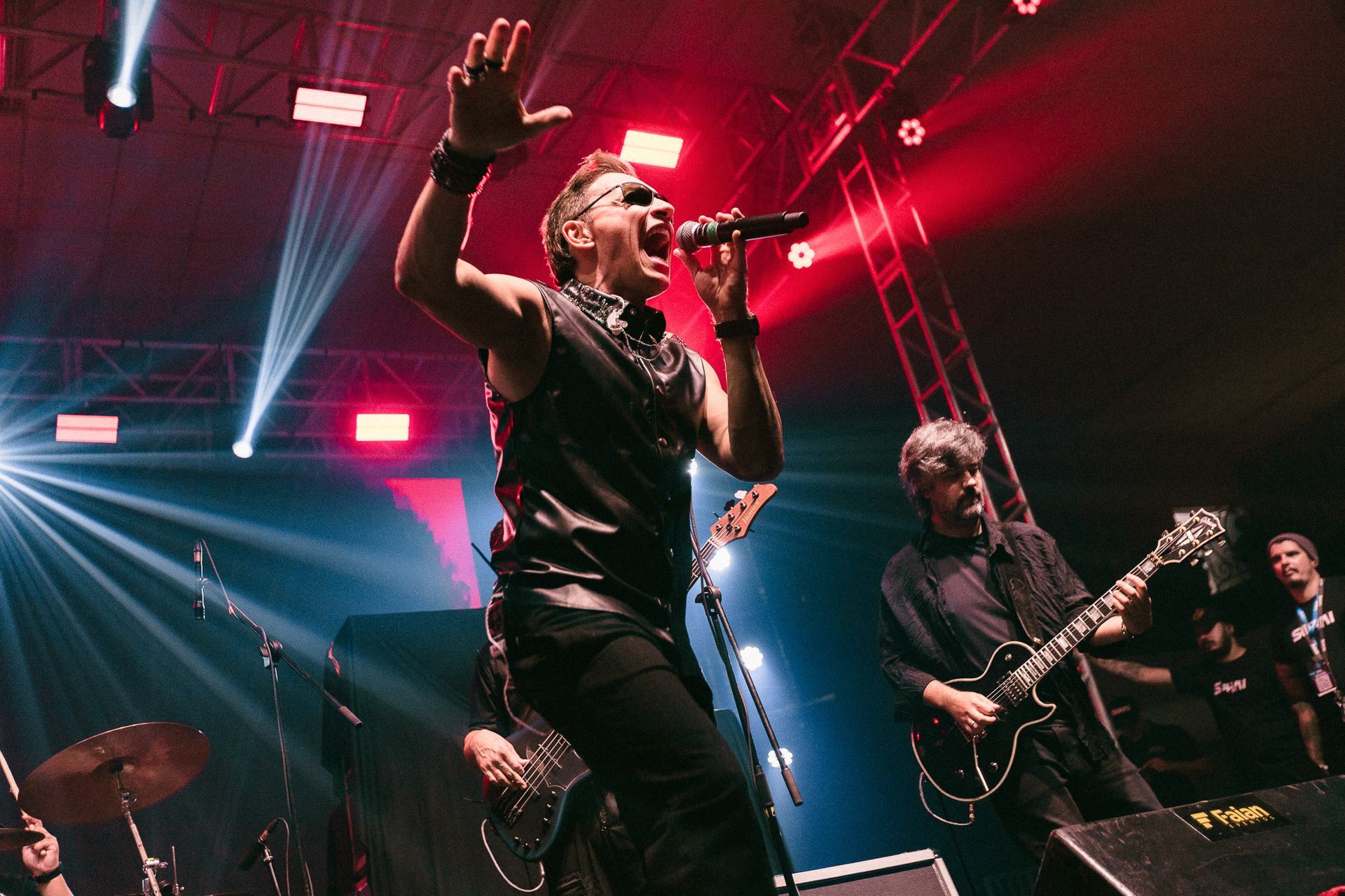 Banda se apresentando no palco com o vocalista segurando o microfone, braços erguidos. Iluminação de palco vermelha e azul.