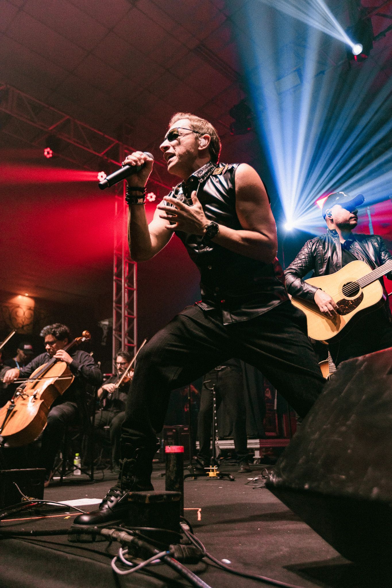 Homem vestido de preto cantando no palco, iluminado por luzes vermelhas e azuis, acompanhado por uma banda.