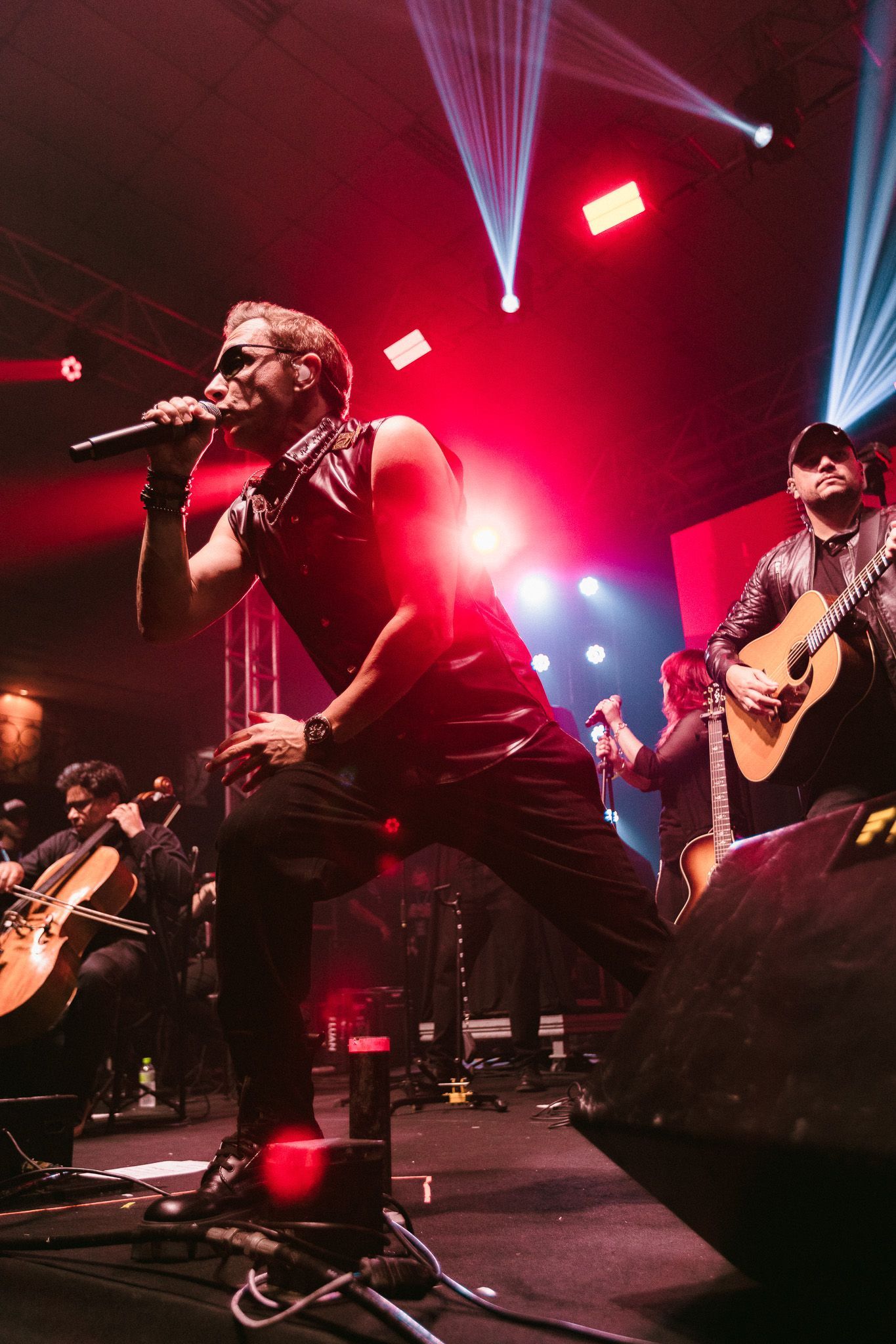 Cantor no palco, cantando ao microfone, com a banda tocando guitarras sob luzes vermelhas e azuis.
