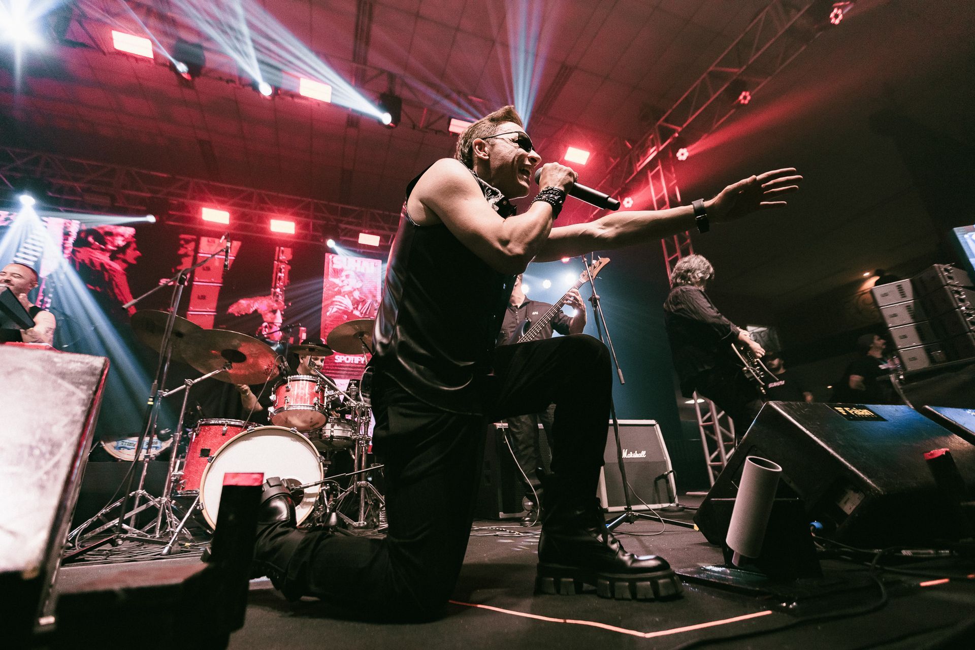 Um cantor se ajoelha, cantando em um microfone. Luzes de palco iluminam uma banda que se apresenta para uma plateia.