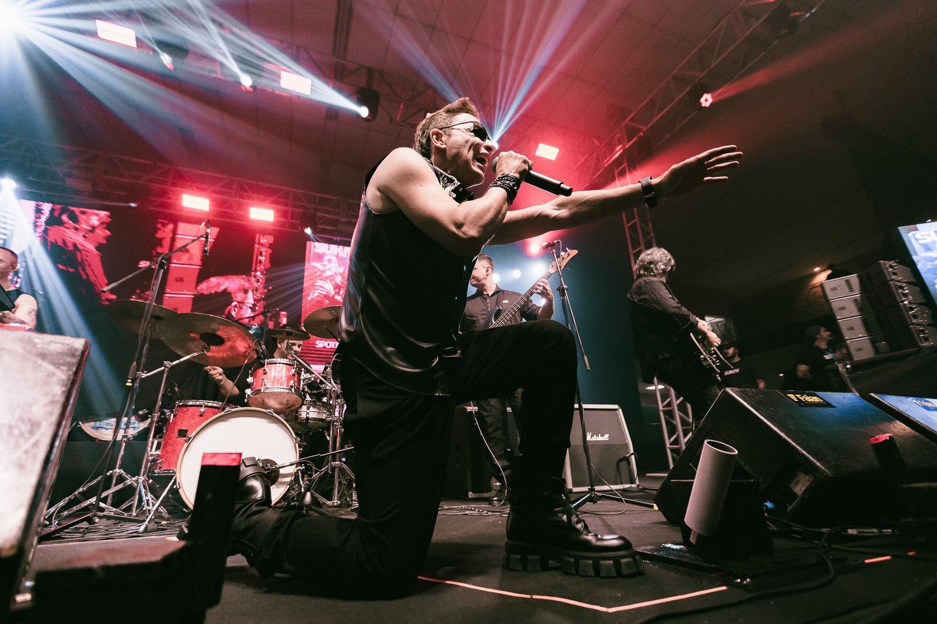 Uma banda se apresenta no palco sob luzes brilhantes, o vocalista se ajoelha e canta em um microfone.