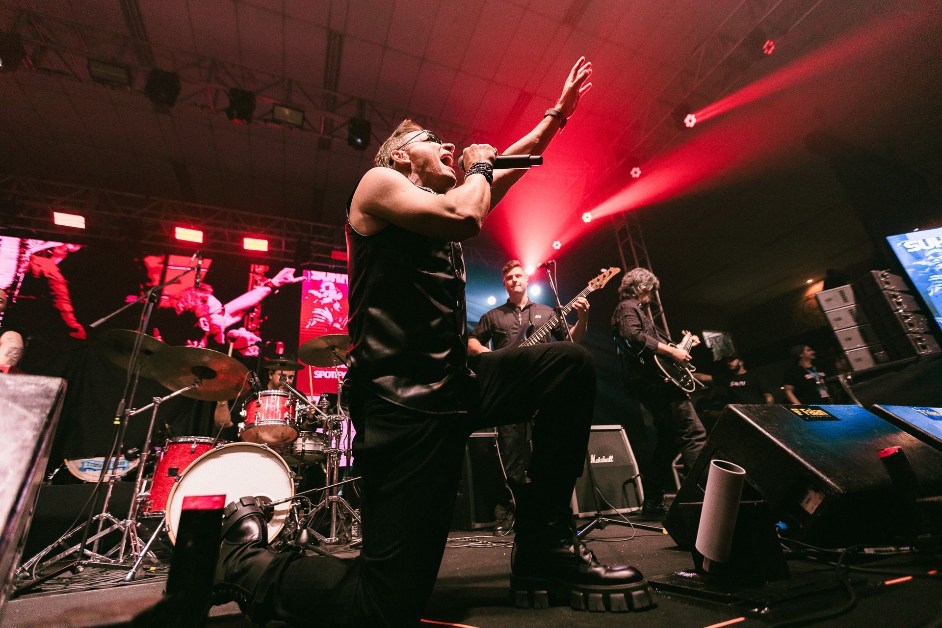 Banda se apresentando no palco com iluminação vermelha. O vocalista principal se ajoelha, com o braço erguido.