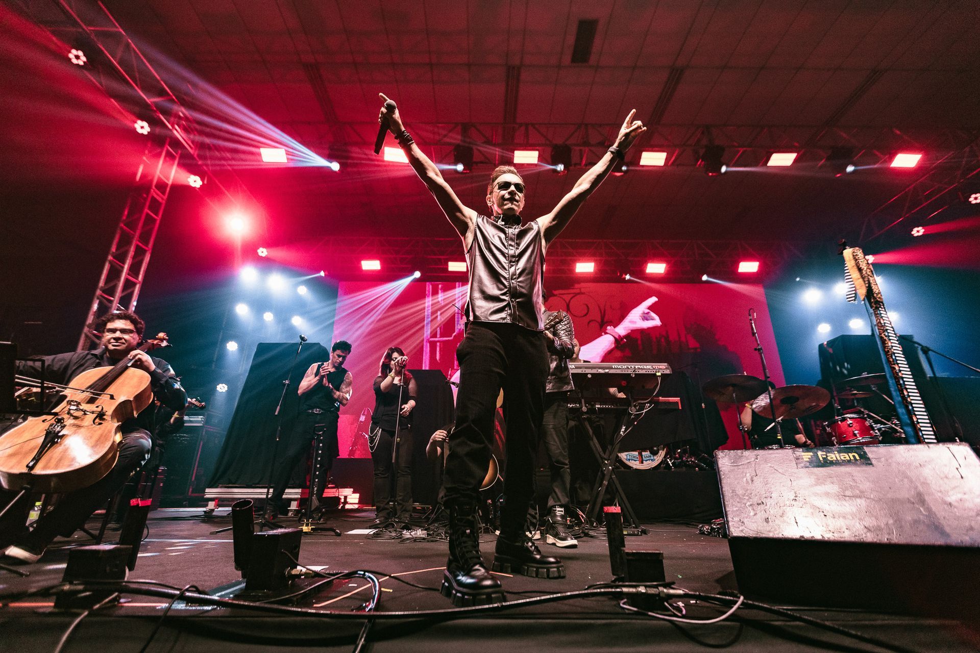 Banda se apresentando no palco, vocalista com os braços erguidos, iluminação de palco vermelha e azul.