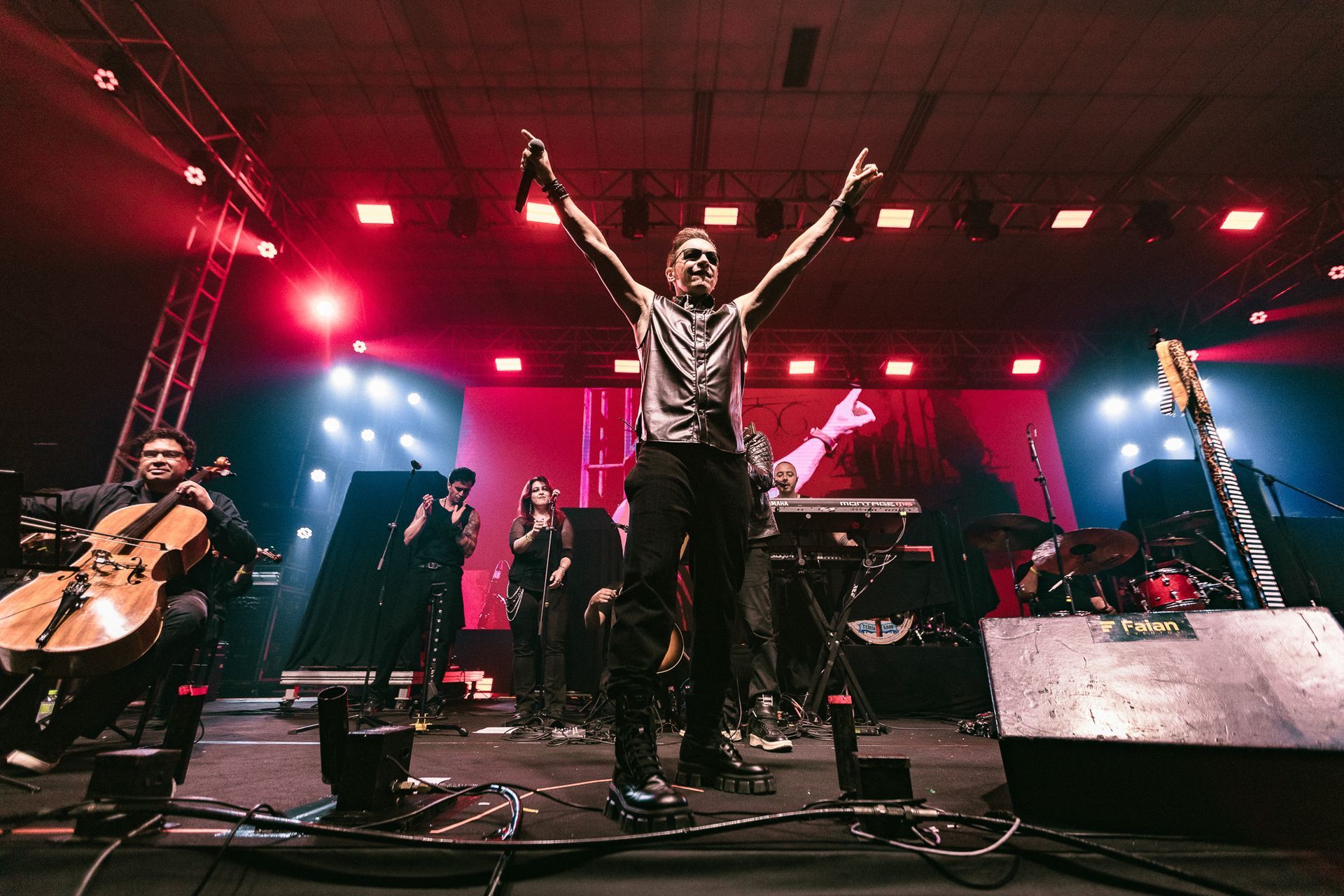 Banda se apresentando no palco com os braços erguidos. A iluminação cênica em vermelho e azul destaca os músicos 