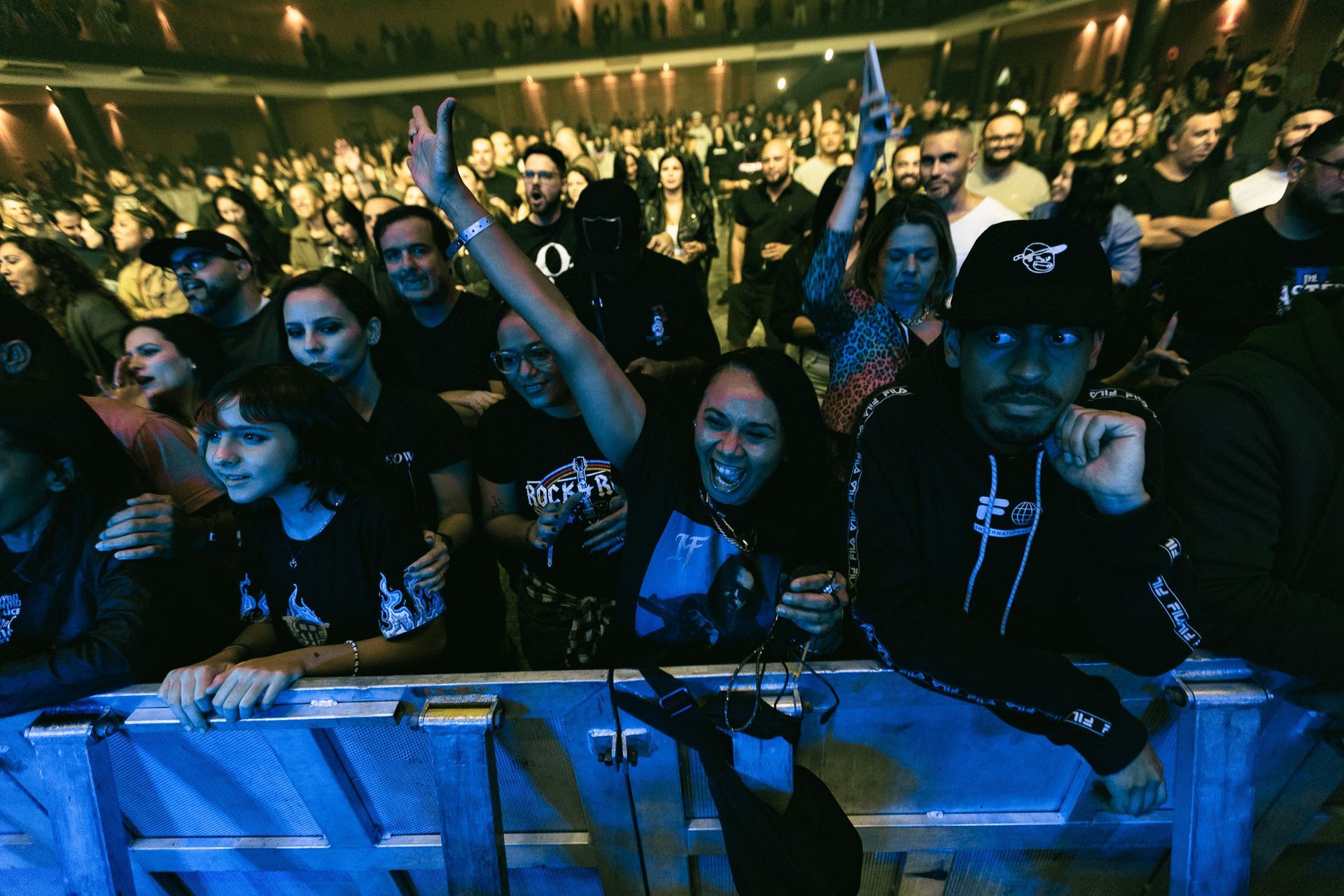 Pessoas em uma plateia de show, algumas aplaudindo, muitas vestindo preto. O palco está fora do enquadramento.