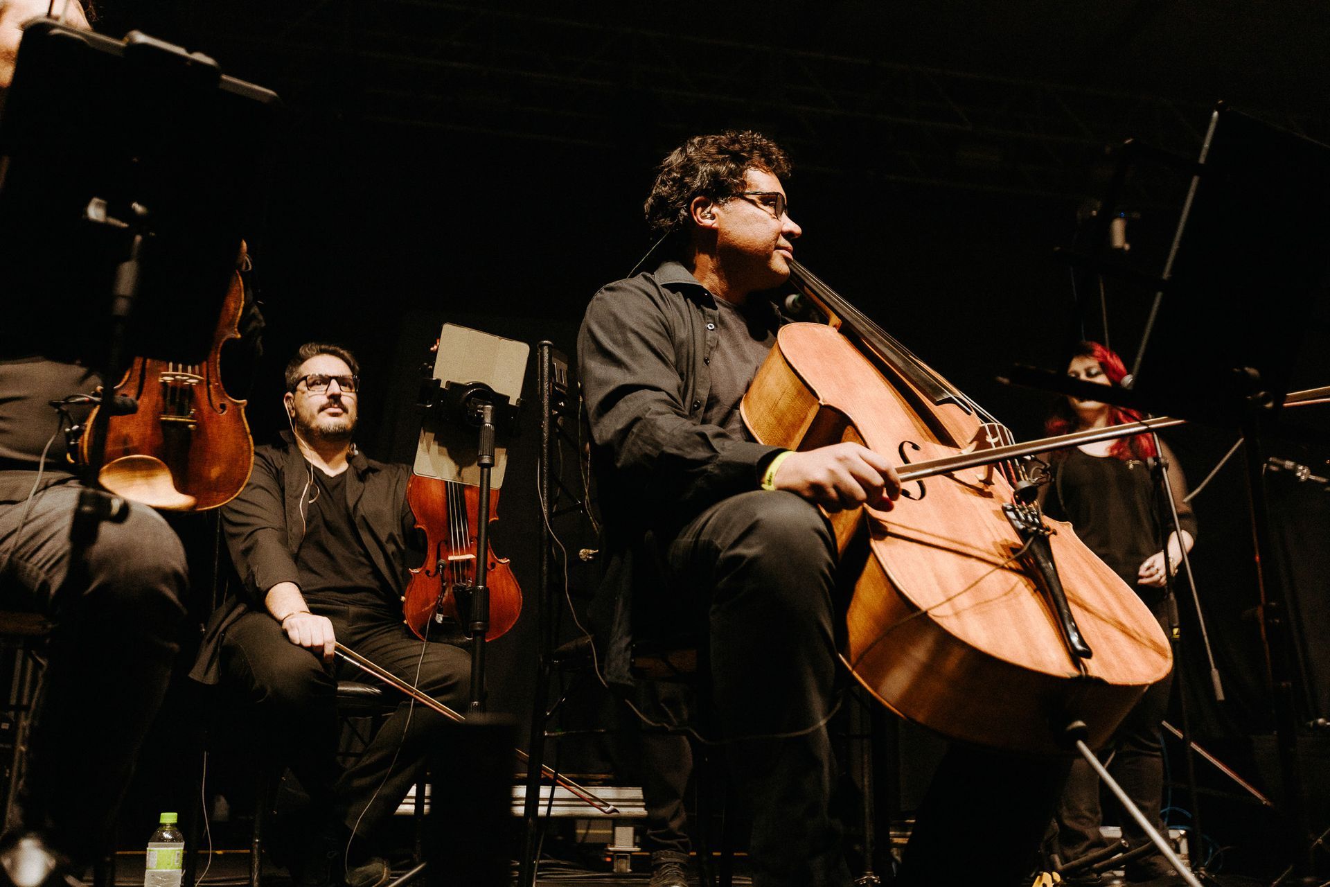 Um músico tocando um violoncelo grande no palco com outros músicos vestidos de preto.