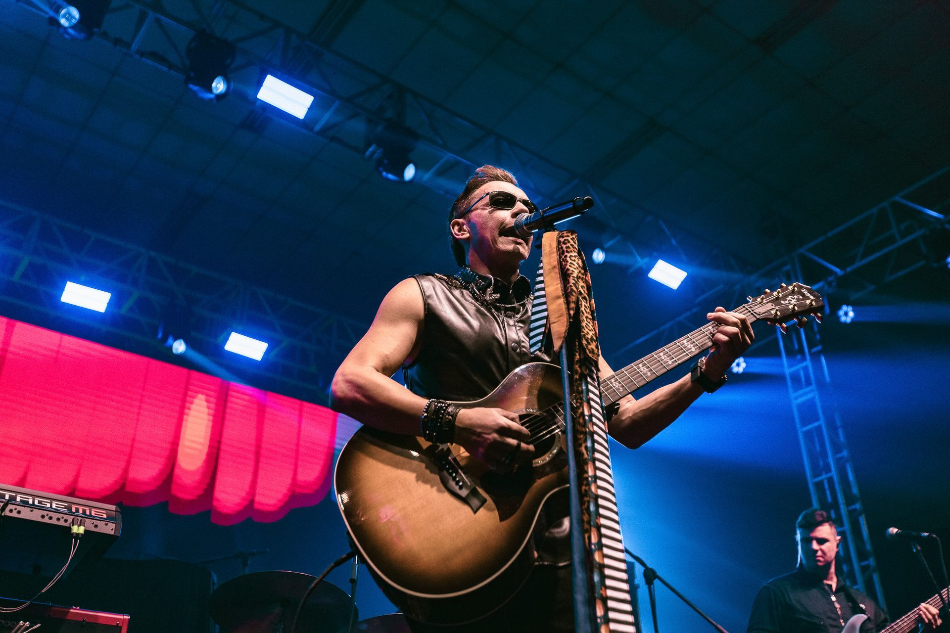 Homem tocando violão no palco, cantando ao microfone. Roupas escuras, luzes de palco.