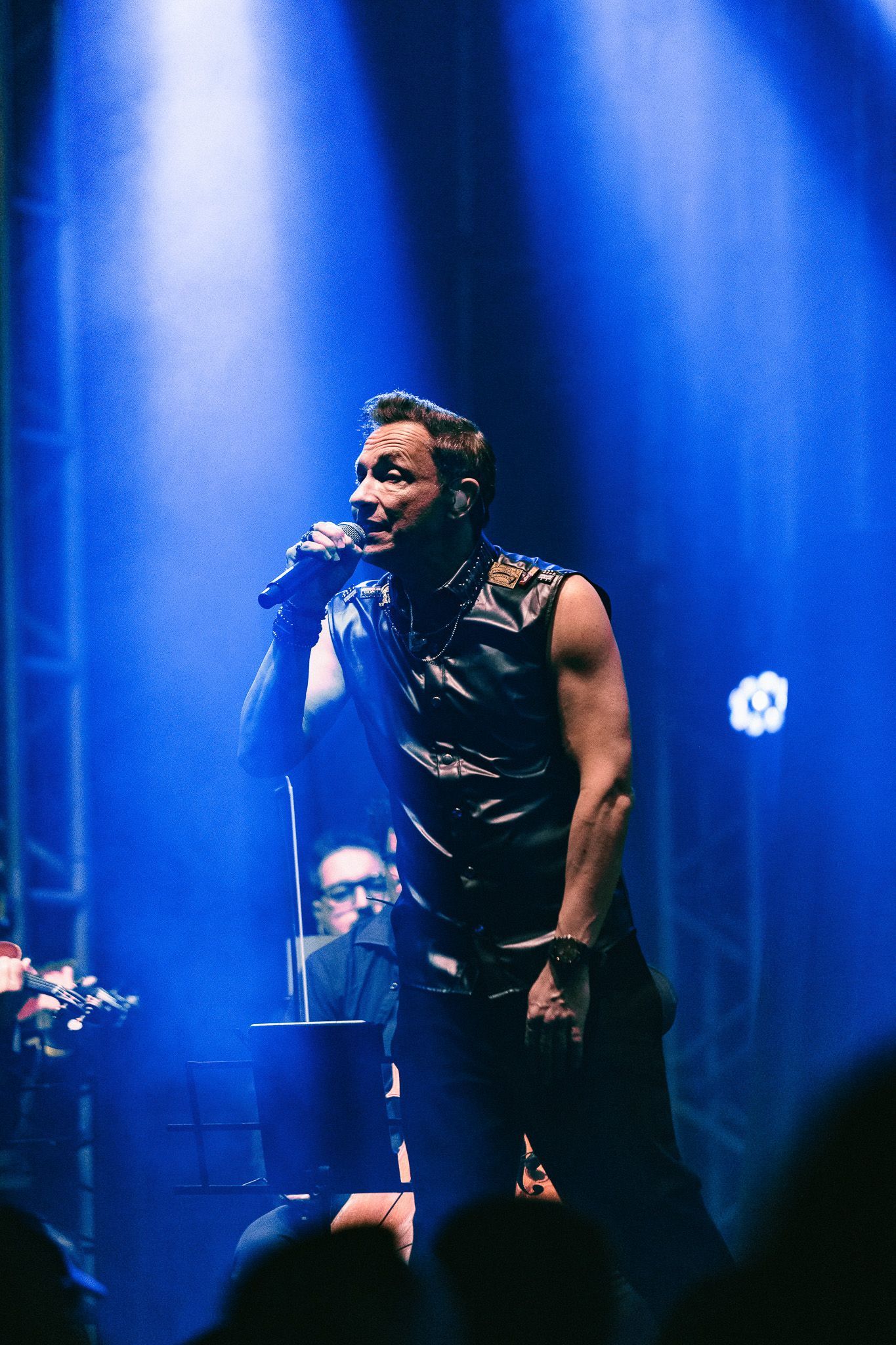 Cantor no palco, iluminada por luzes azuis, segurando um microfone e cantando.