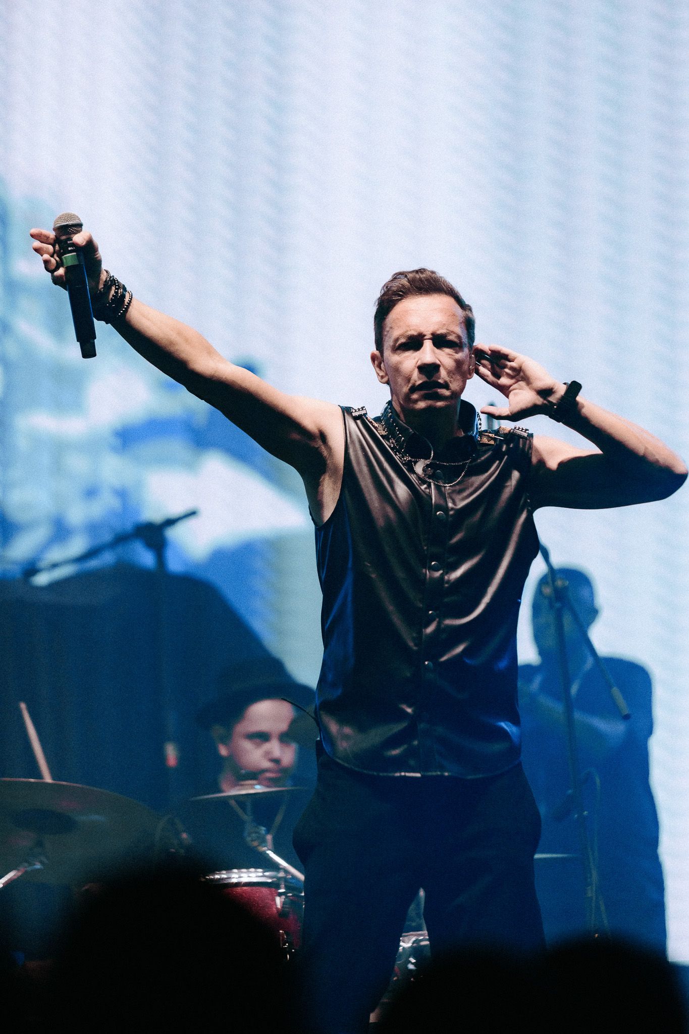 Cantor no palco com microfone, braço levantado, vestindo camisa sem mangas. Banda ao fundo