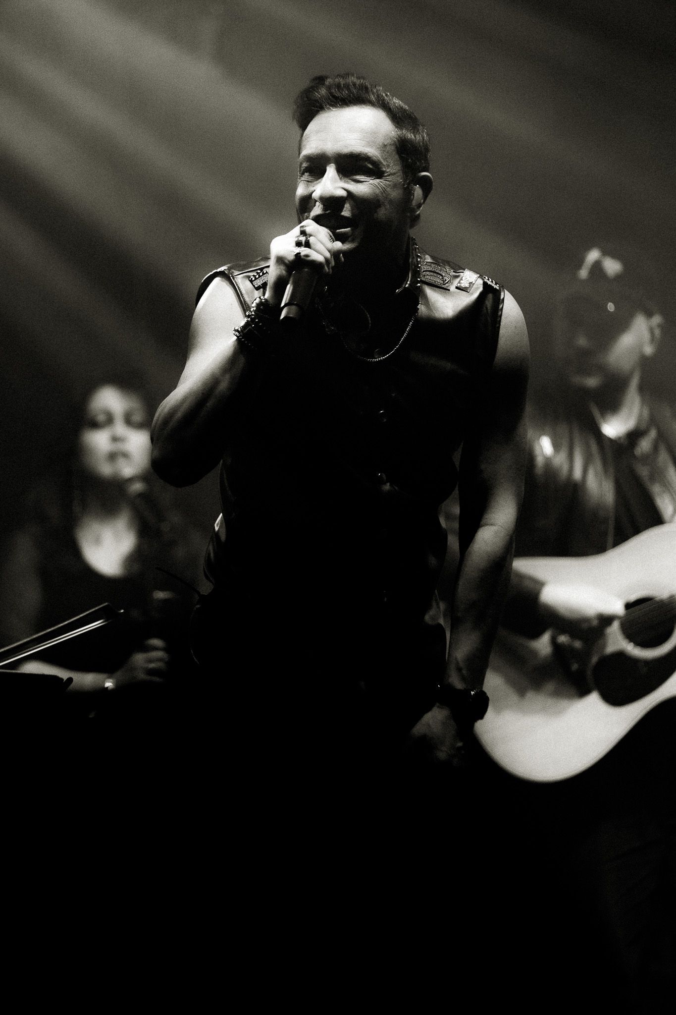 Cantor com microfone no palco, sorrindo; colegas de banda ao fundo, tocando instrumentos. Preto e branco.