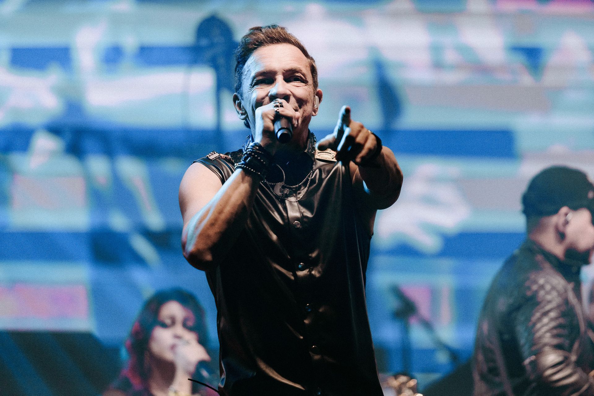 Homem cantando ao microfone no palco, apontando para a frente. Integrantes da banda ao fundo.