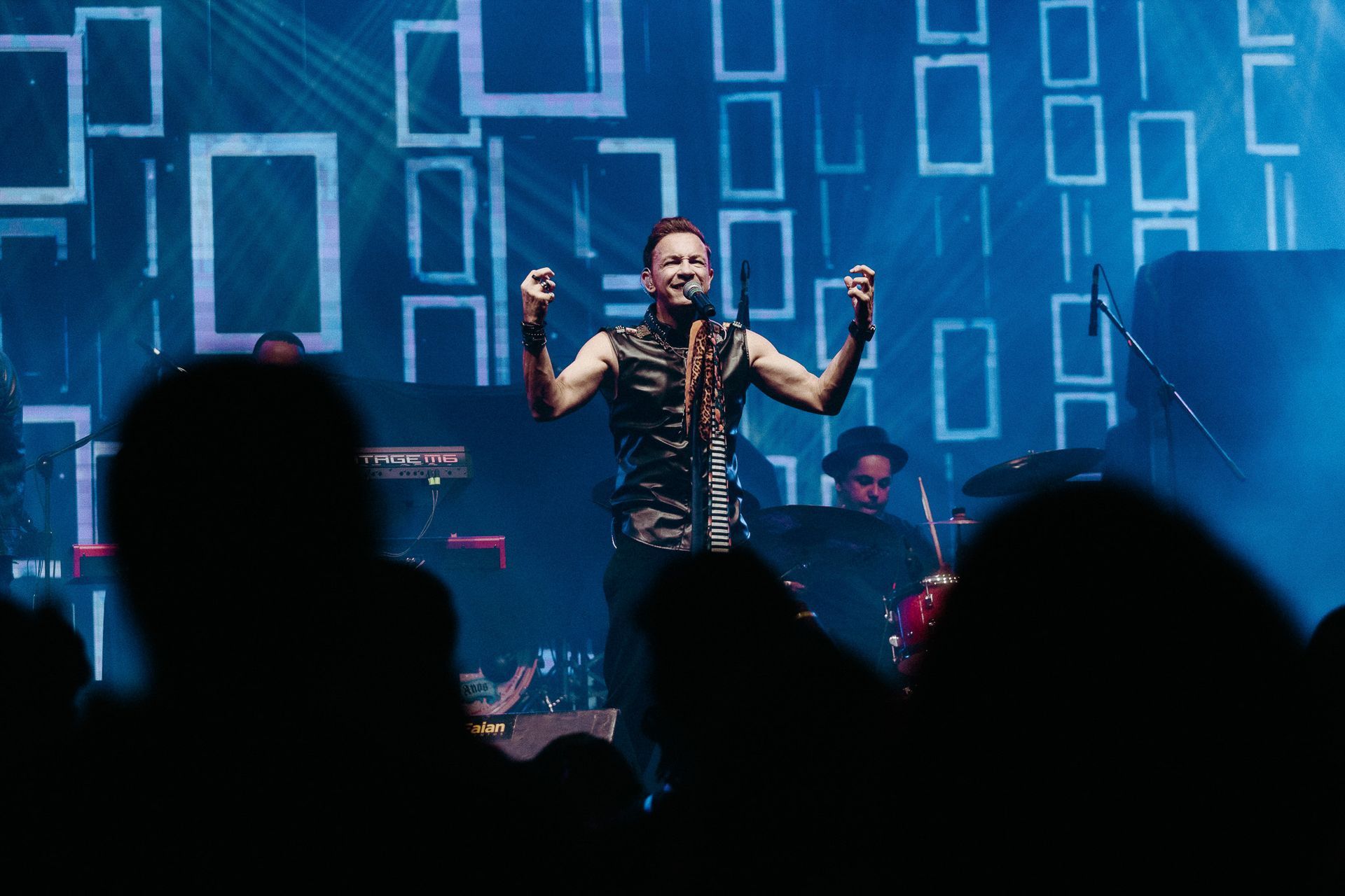 Cantor se apresentando no palco com os braços erguidos, banda ao fundo. Palco iluminado com luz azul e padrões quadrados.