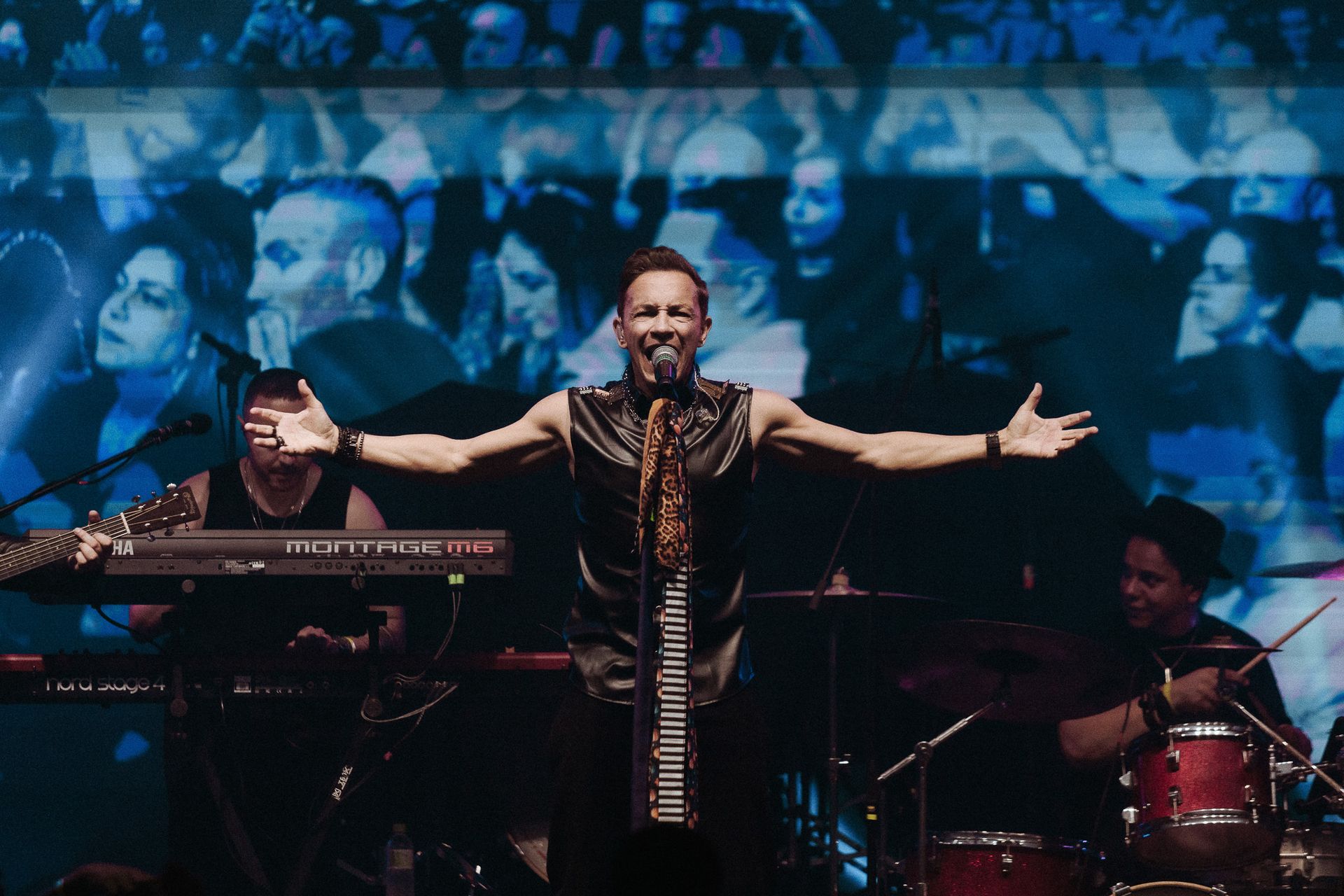 Uma banda se apresentando no palco. Vocalista com os braços estendidos.
