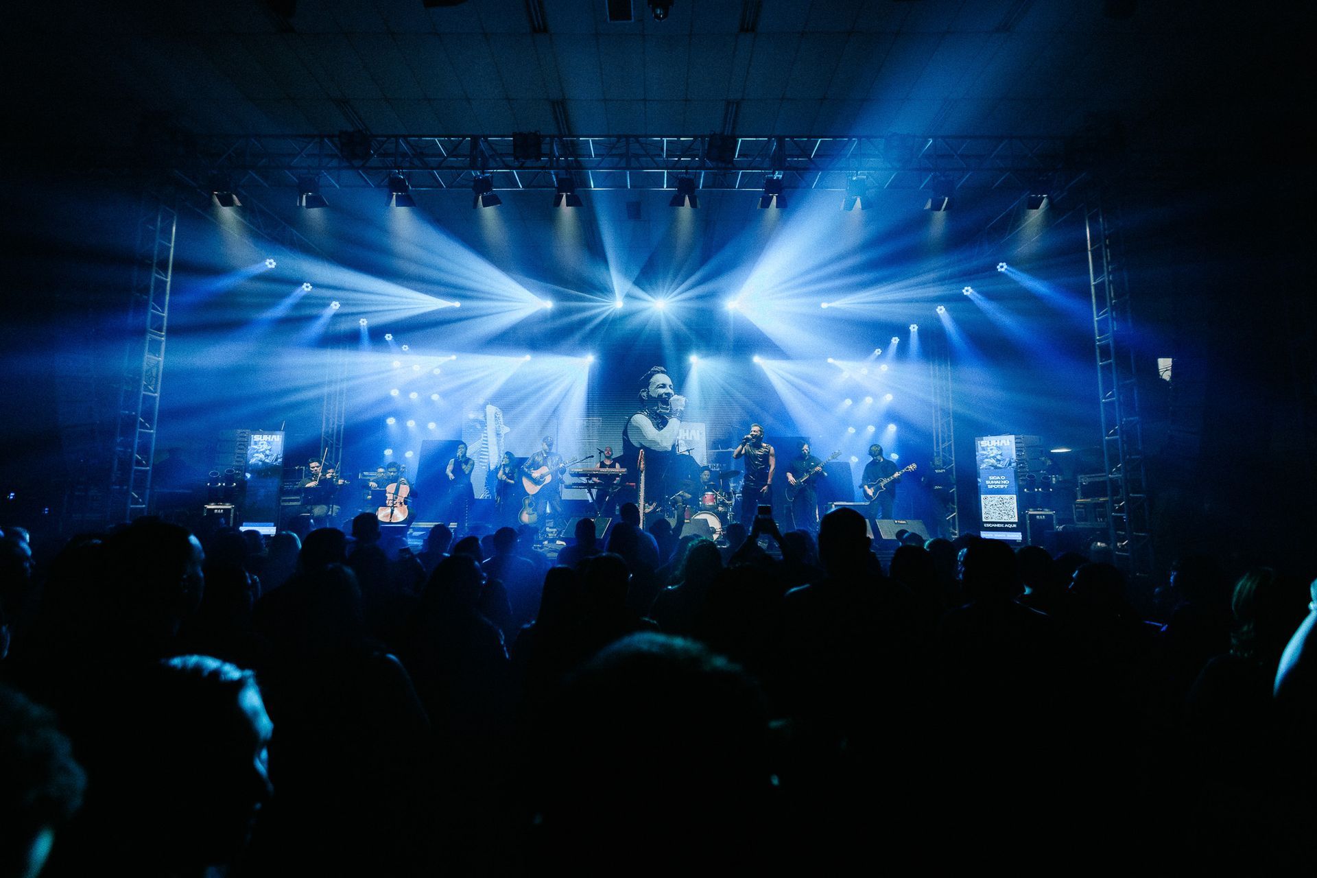 Palco de concertos com holofotes azuis brilhantes iluminando a banda e o público em um local escuro.