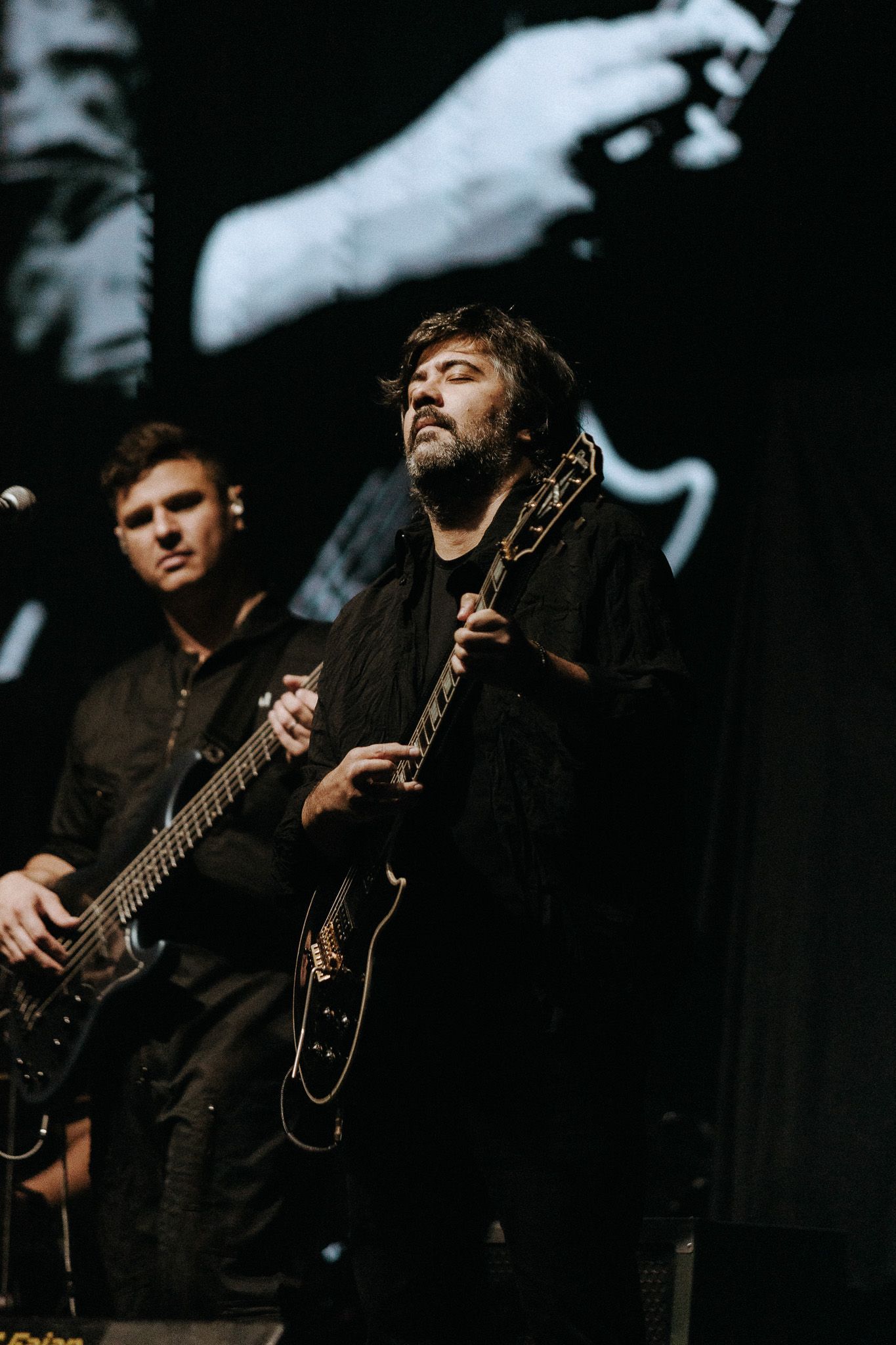 Dois músicos no palco, um tocando guitarra e o outro baixo. Ambiente escuro com fundo projetado.
