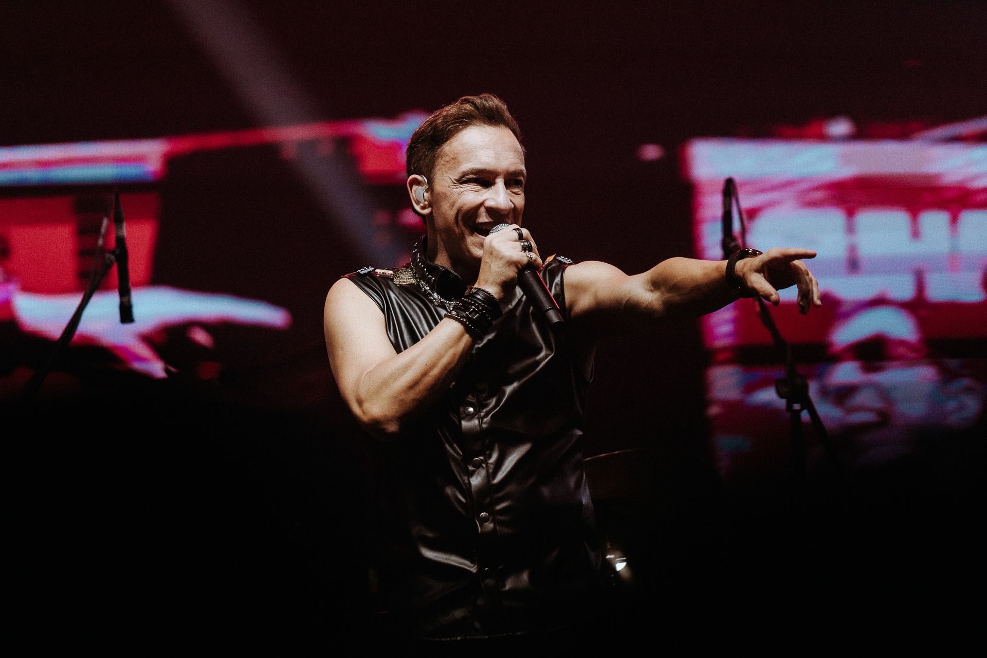 Um artista cantando no palco, apontando com um dos braços. Ele veste um colete de couro preto e segura um microfone.