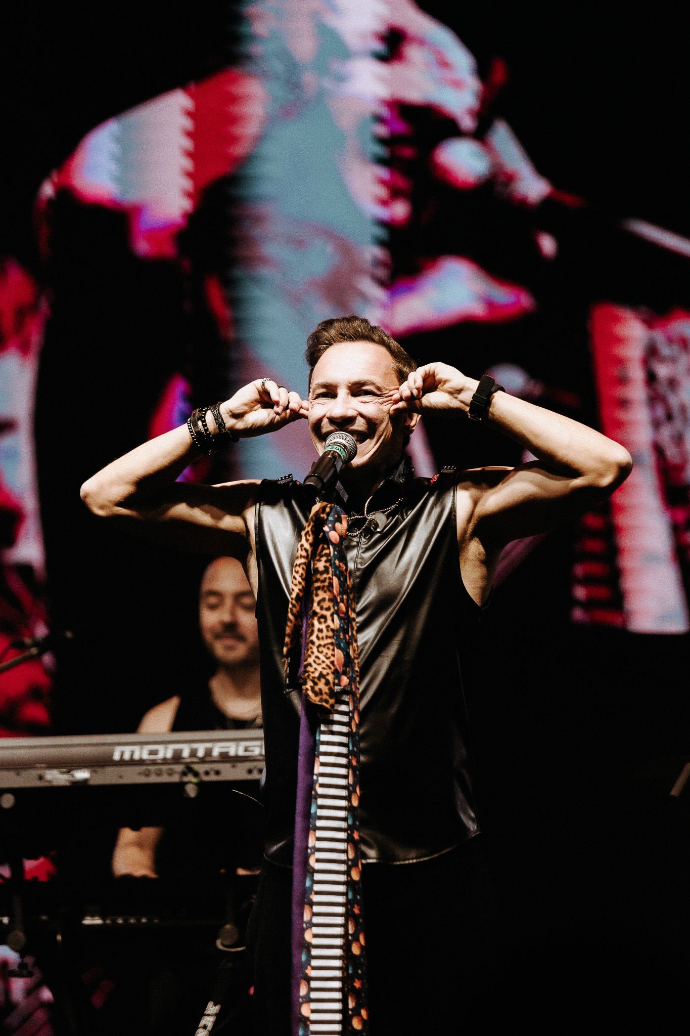 Cantor no palco com as mãos perto do rosto, sorrindo. Pessoa ao teclado ao fundo.