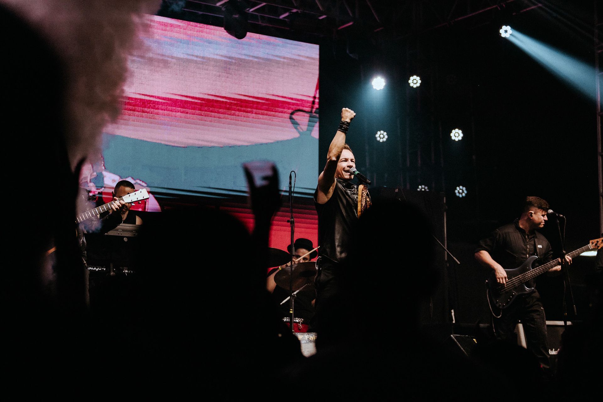 Banda se apresentando no palco, vocalista com o punho erguido, luzes brilhantes, público em primeiro plano.