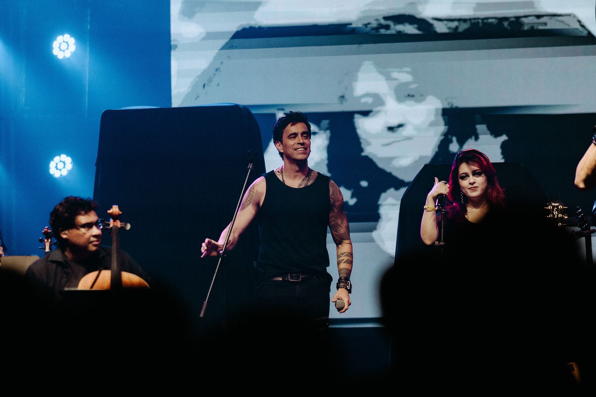 Uma banda se apresenta no palco. Um homem de regata sorri enquanto gesticula. Uma cantora de cabelo rosa