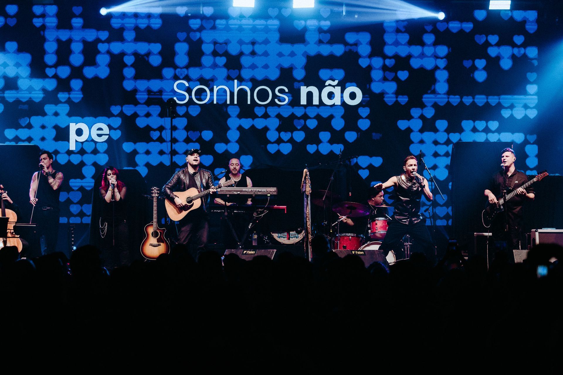 Banda se apresentando no palco com fundo azul, com a frase 