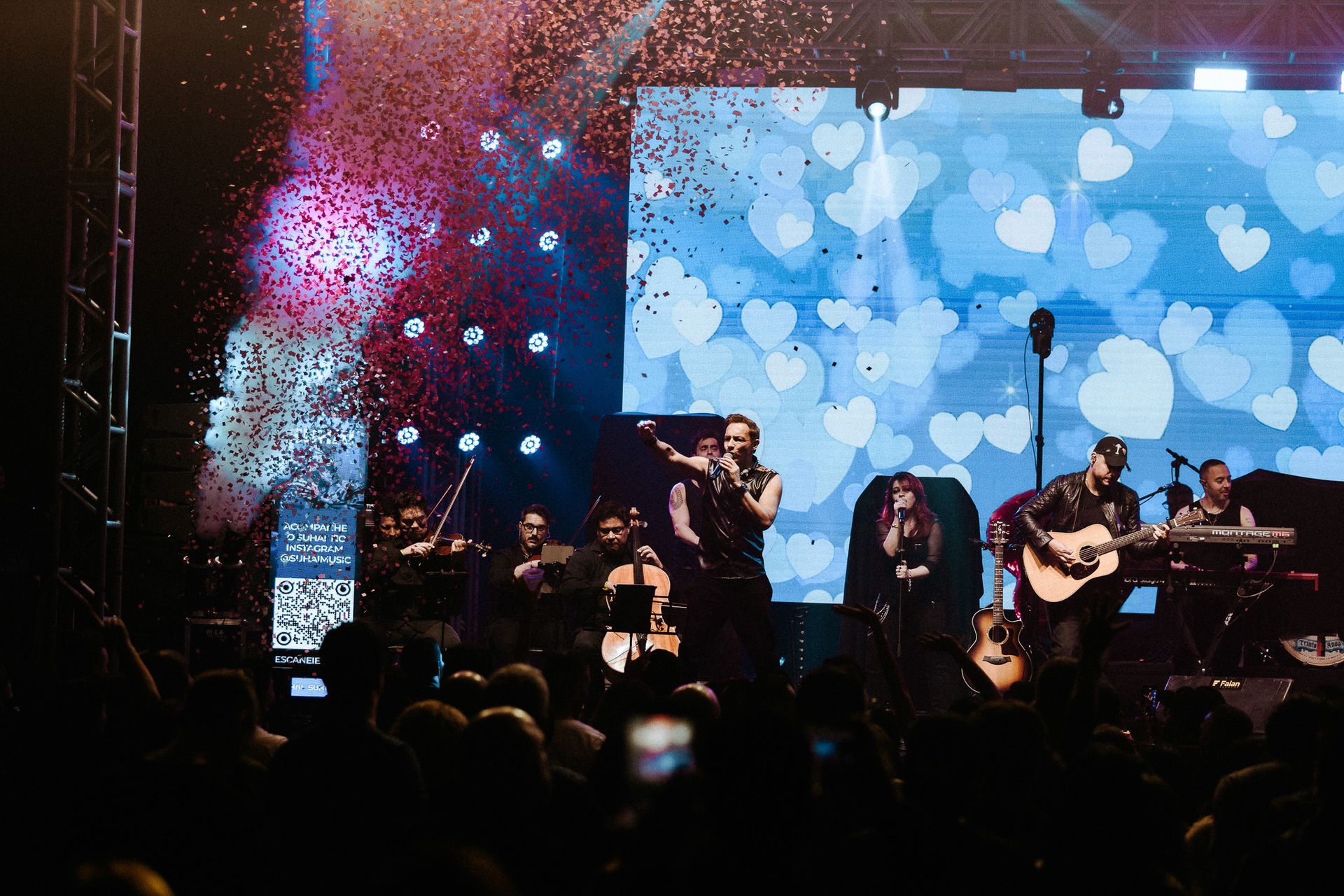 Banda se apresentando no palco com confetes caindo em frente a uma tela colorida.